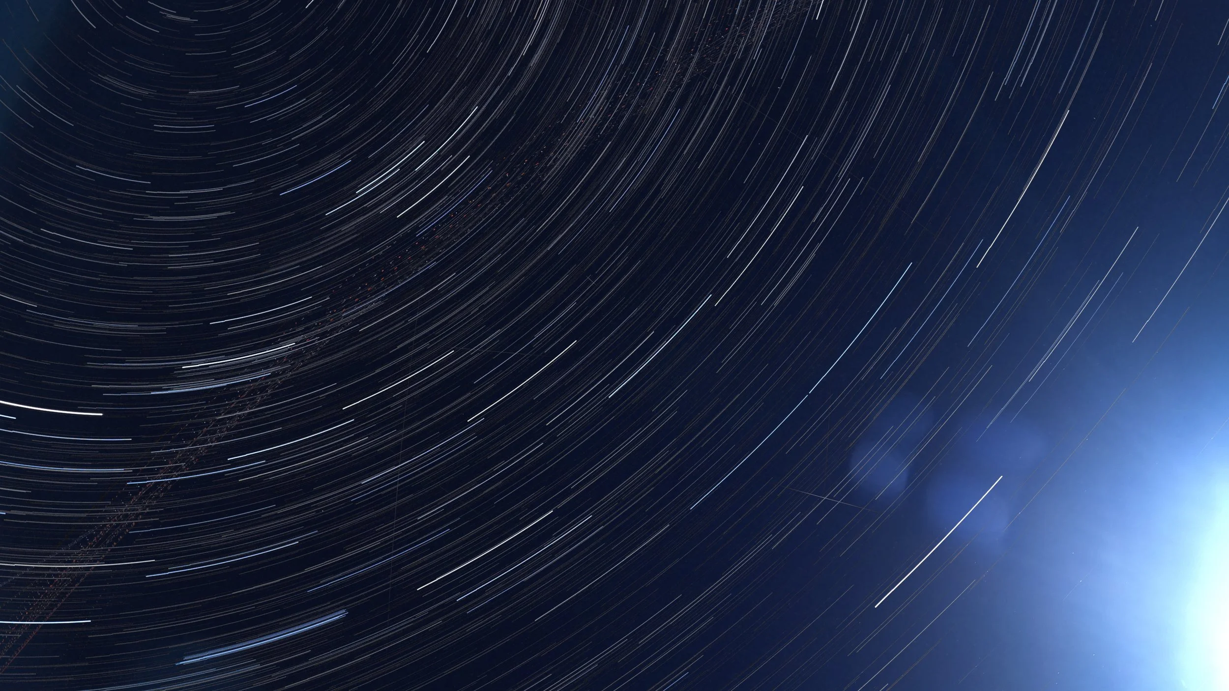Long exposure photo of star trails in the night sky with a bright light on the horizon.