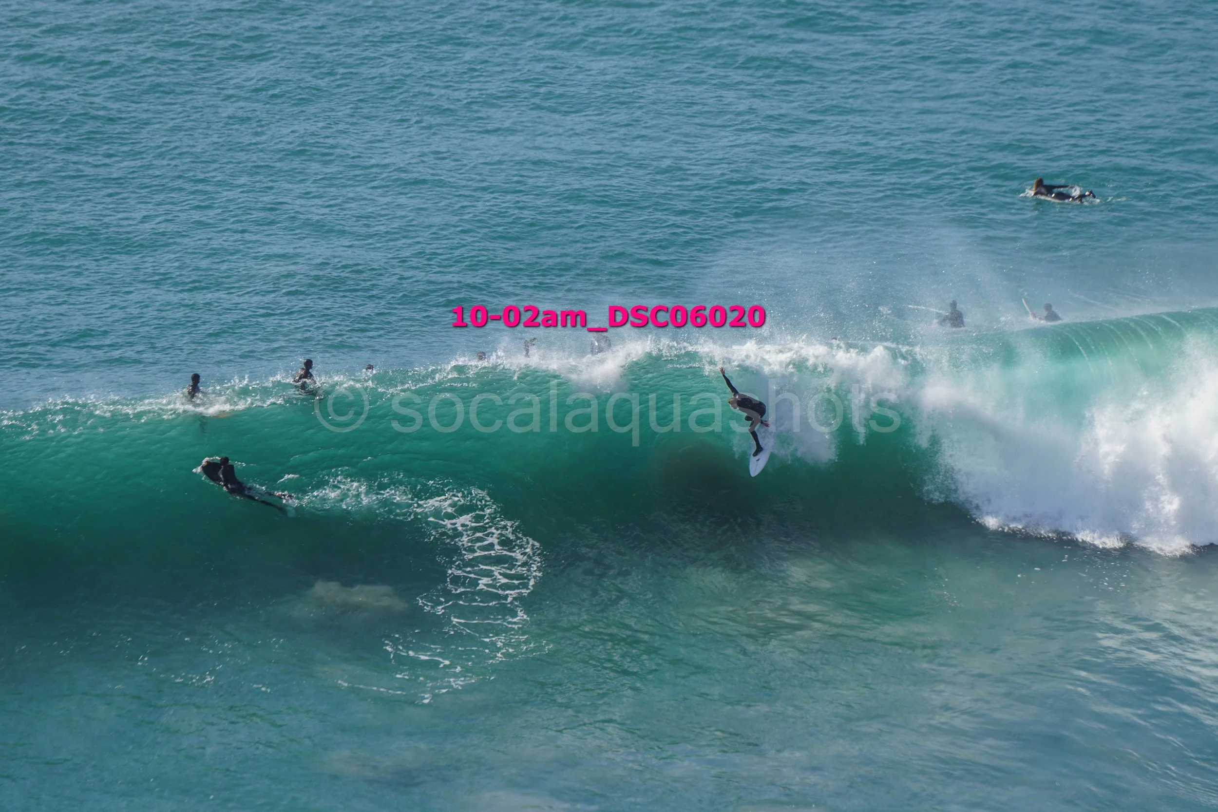 Surfer riding a large wave with several other surfers and swimmers in the water around.