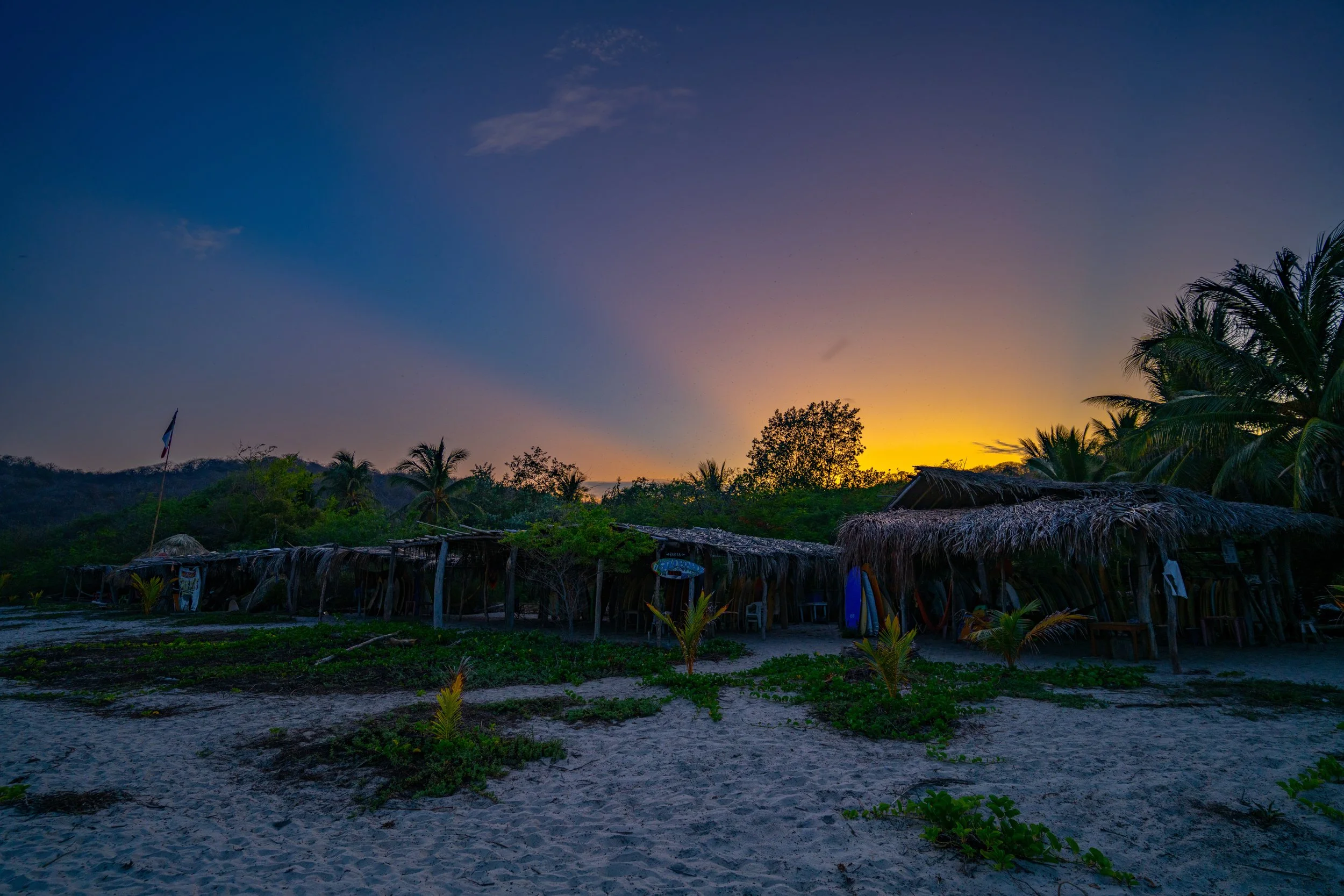 A beach scene at sunset with silhouettes of palm trees, thatched-roof huts, and a flag, under a partly cloudy sky transitioning from blue to orange.