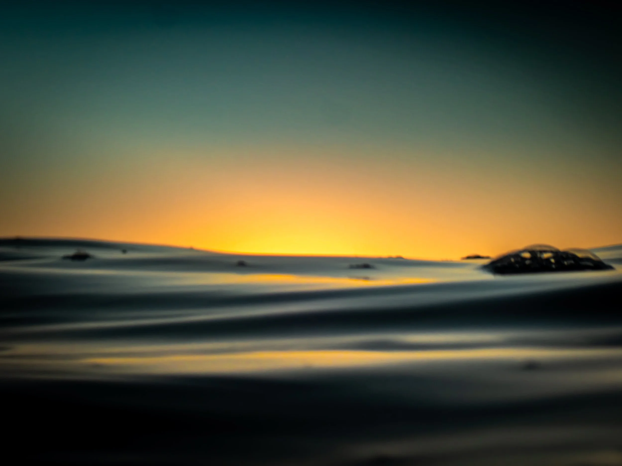 A sunset over calm ocean water with rocks visible on the horizon, casting a warm glow across the water's surface.