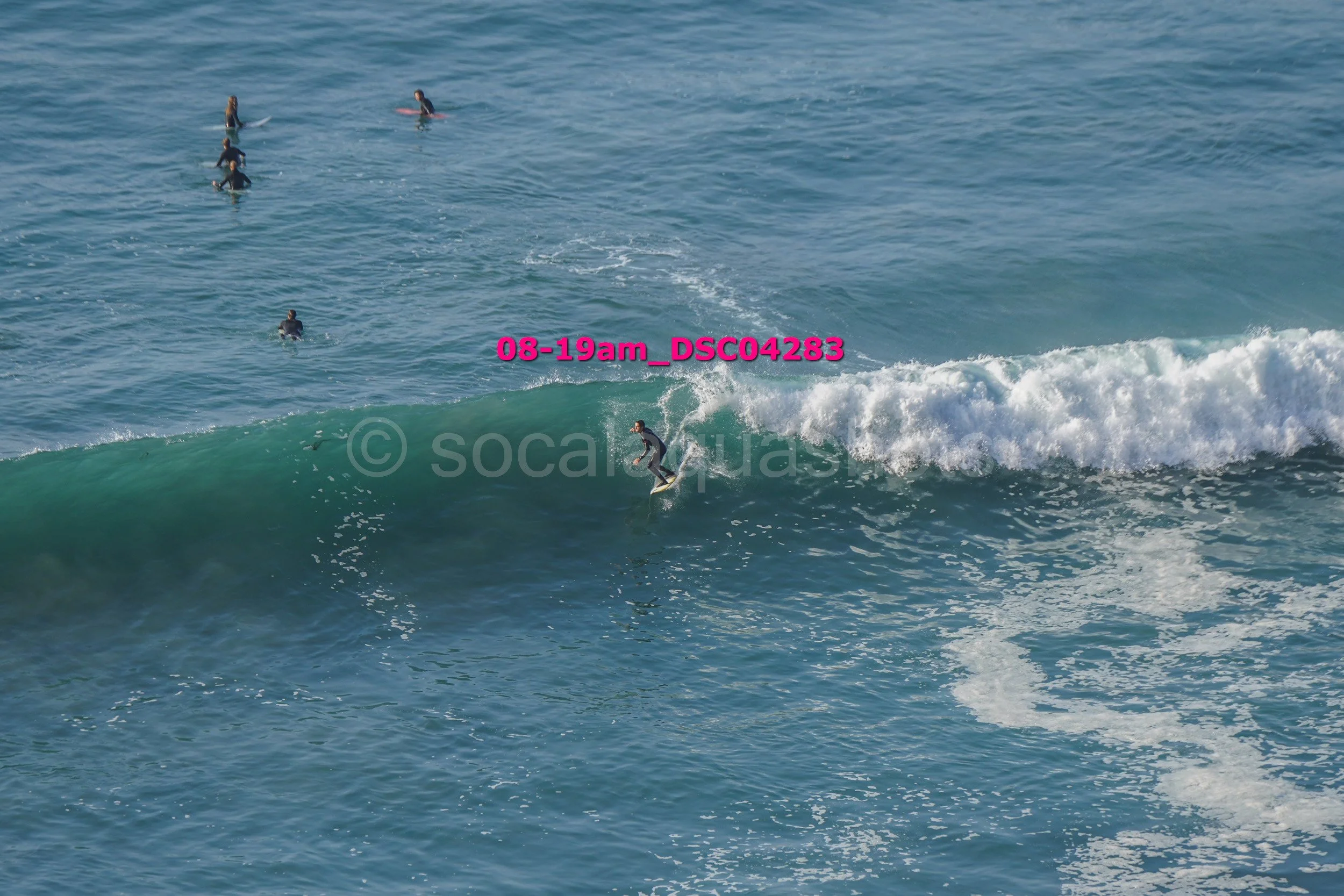 A person surfing on a wave in the ocean while several other surfers wait in the water nearby.