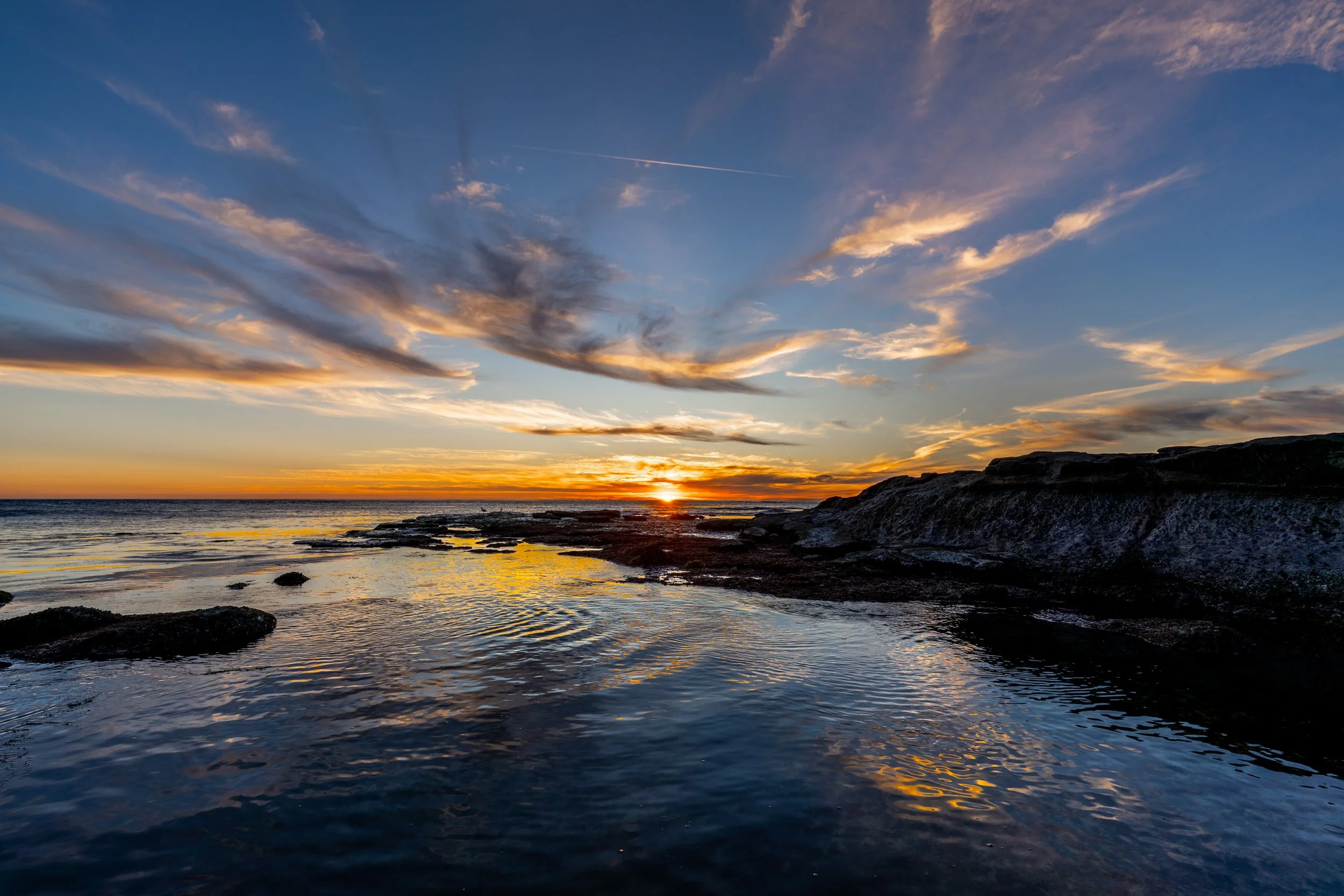 Sunset over the ocean with clouds in the sky and water reflections on rocks in the foreground.