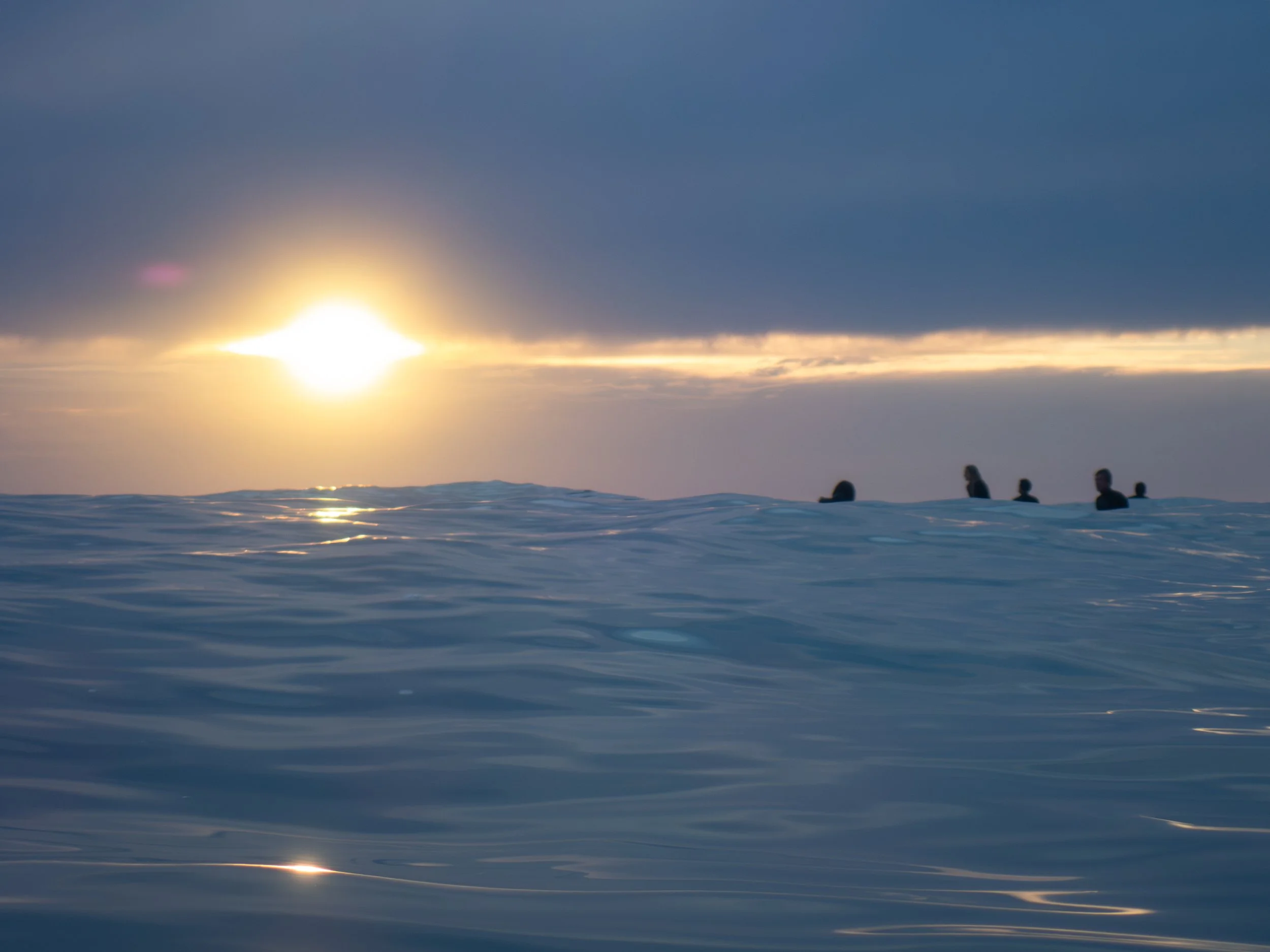 A group of five people swimming or floating in the ocean during sunset, with dark clouds overhead and the sun near the horizon casting a reflection on the water.