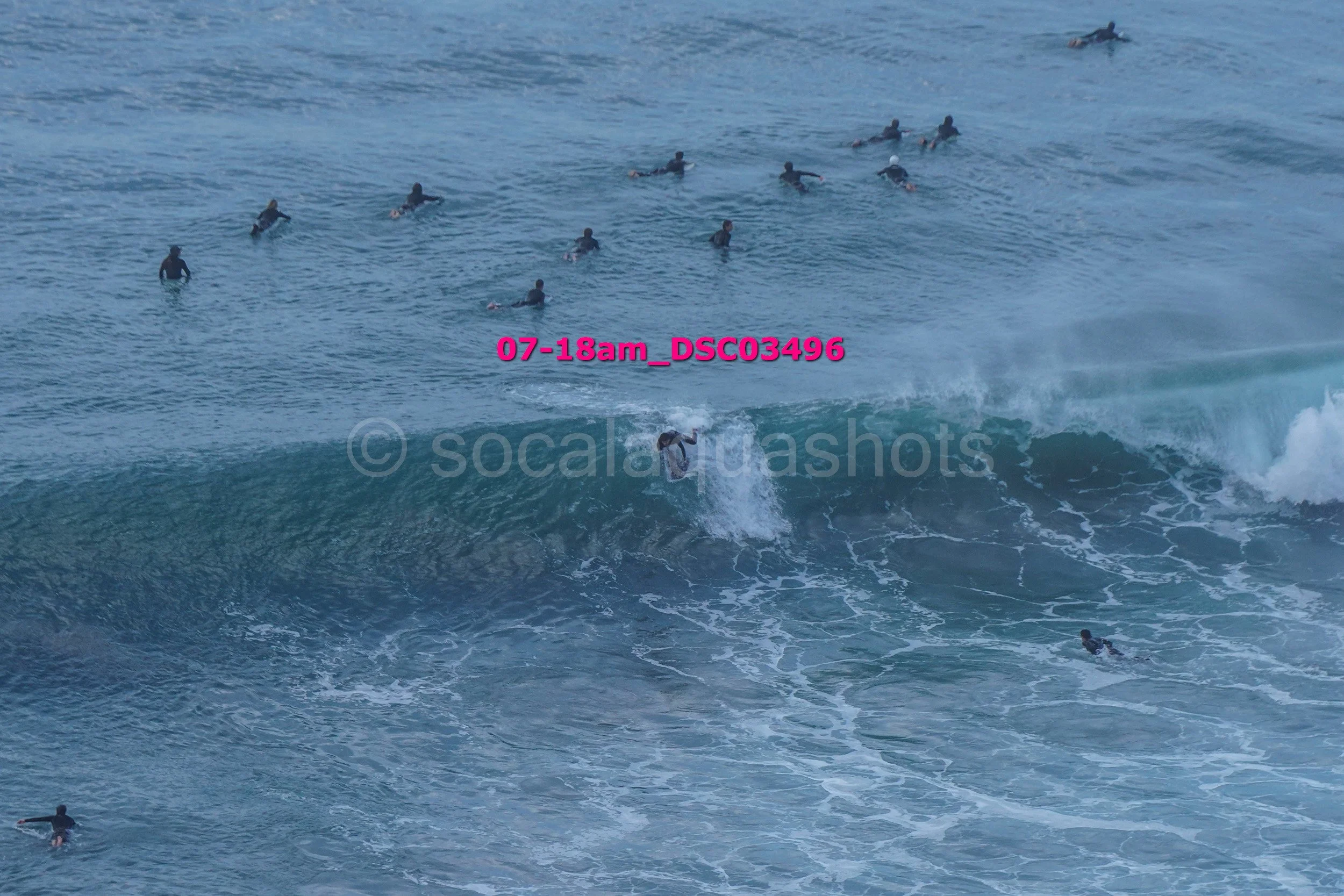 A group of surfers in the ocean, with one surfer riding a wave while others paddle or wait in the water, and the timestamp '07-18am_DSC3496' overlaid in pink text.