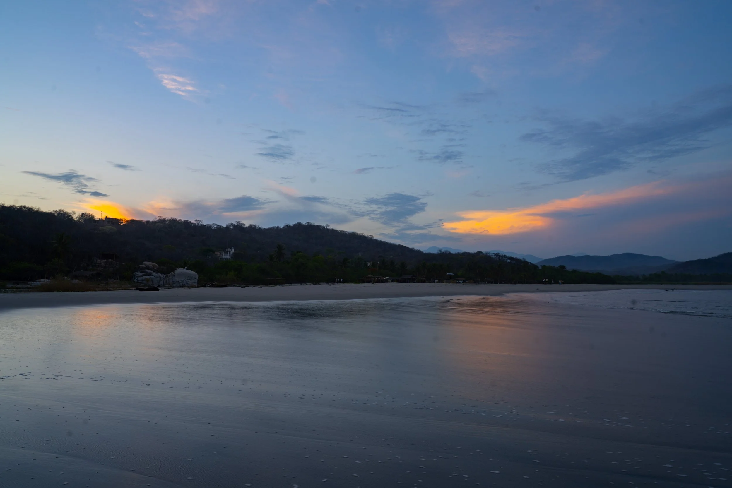 A scenic sunset over a calm beach with a silhouette of a forested hillside and distant mountains.