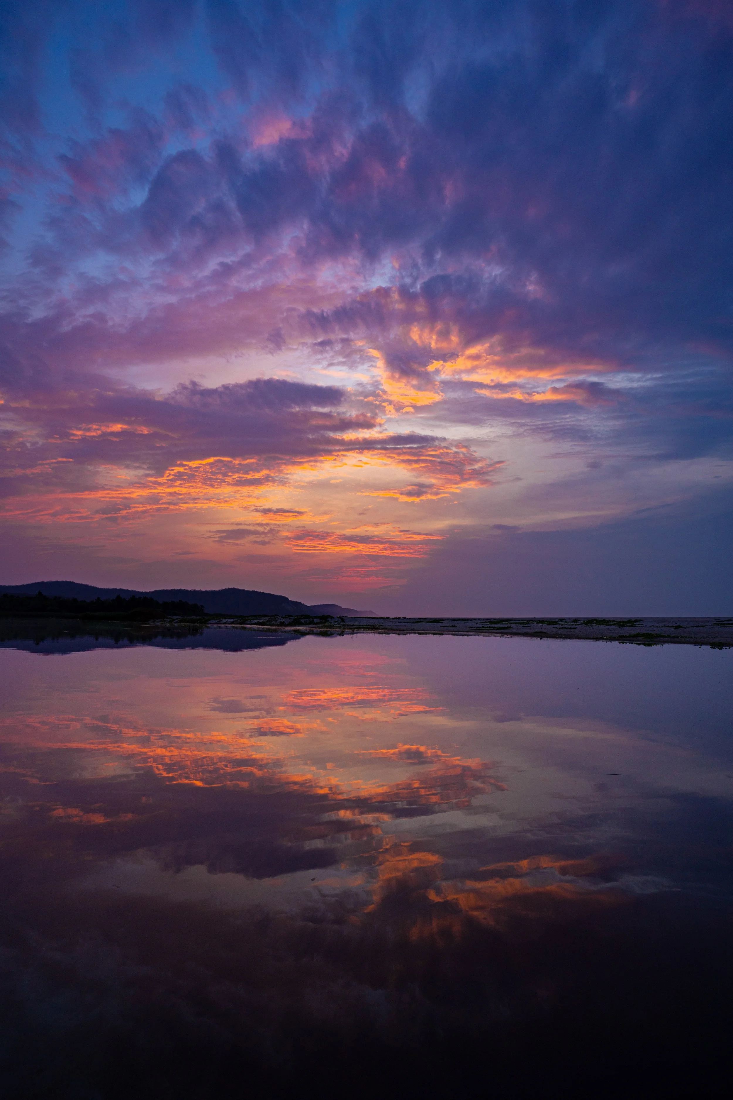 Sunset sky with colorful clouds, purple and orange hues, reflected on a calm body of water with hills in the background.