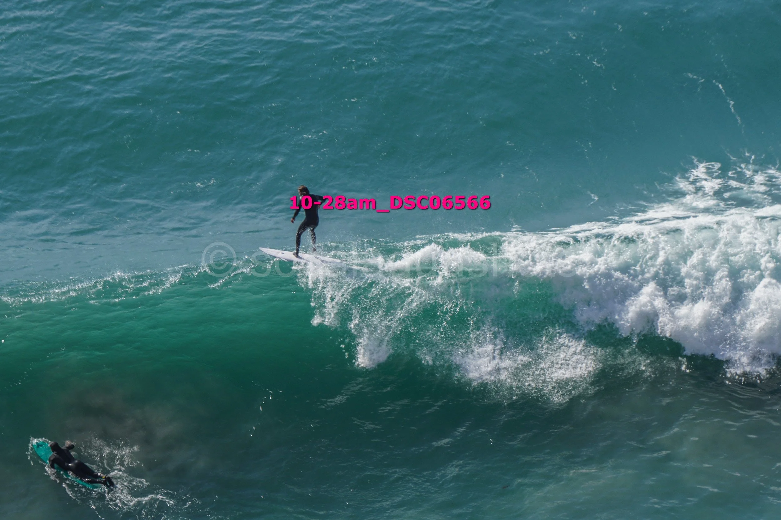 A person surfing on a wave in the ocean, wearing a wetsuit, with another surfer visible in the water below.