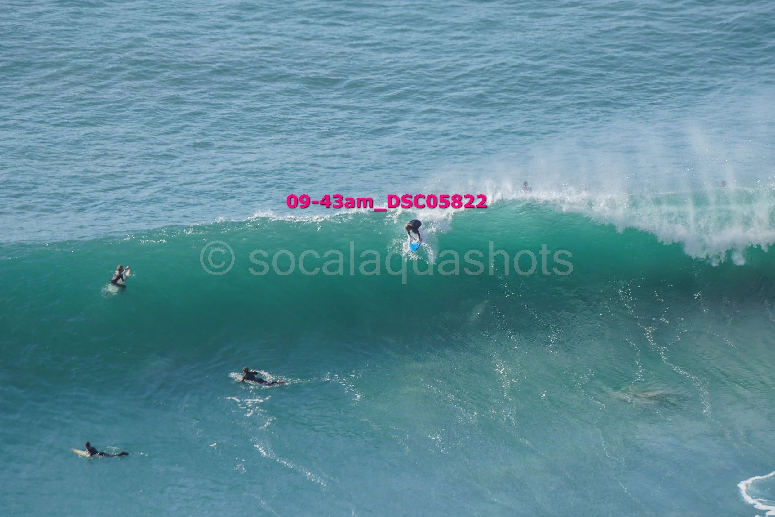 A person surfing on a large wave with several other surfers in the water nearby, ocean in the background.