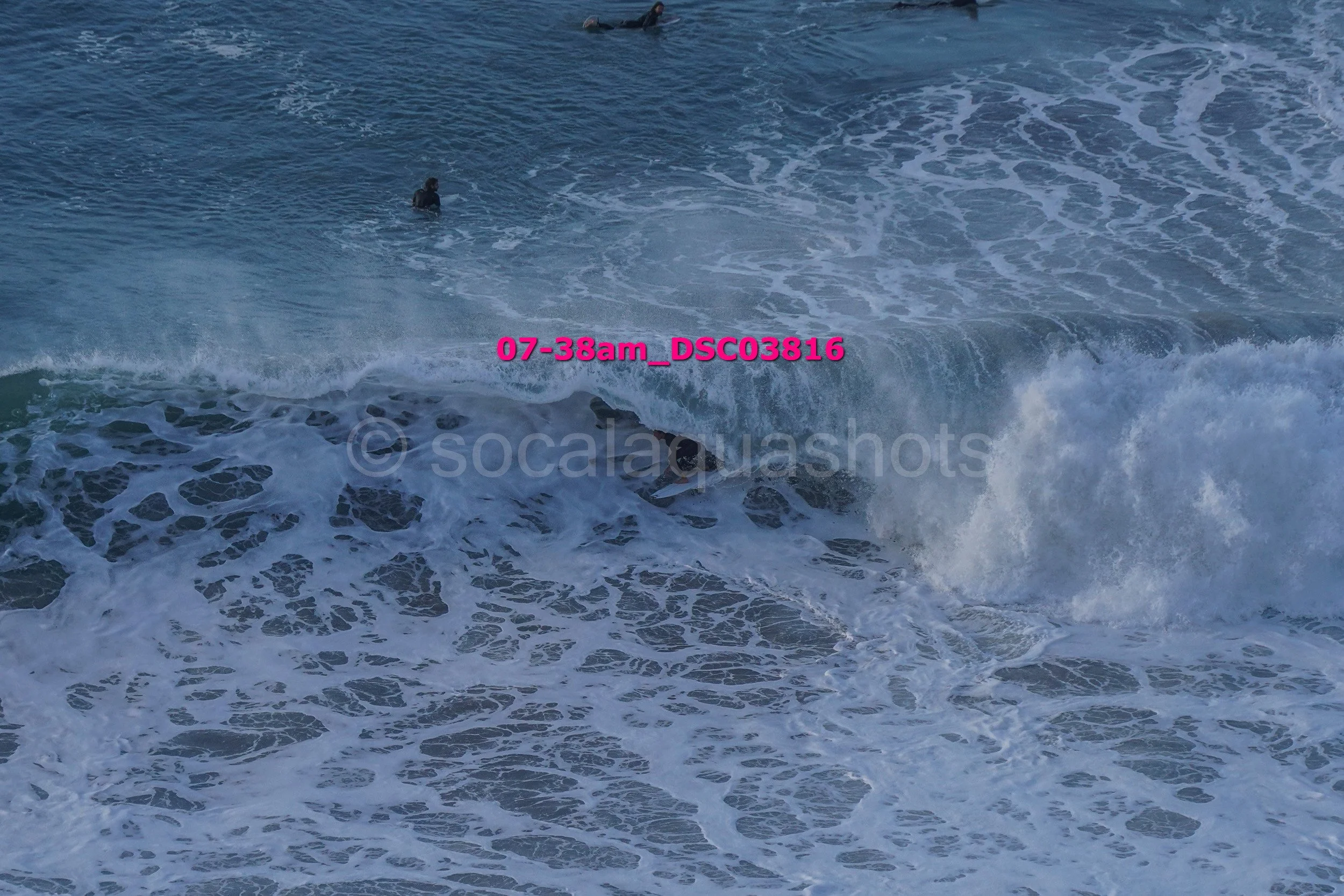 Ocean waves crashing with a surfer partially visible in the water and another person further out, ocean scene at sunrise or sunset.