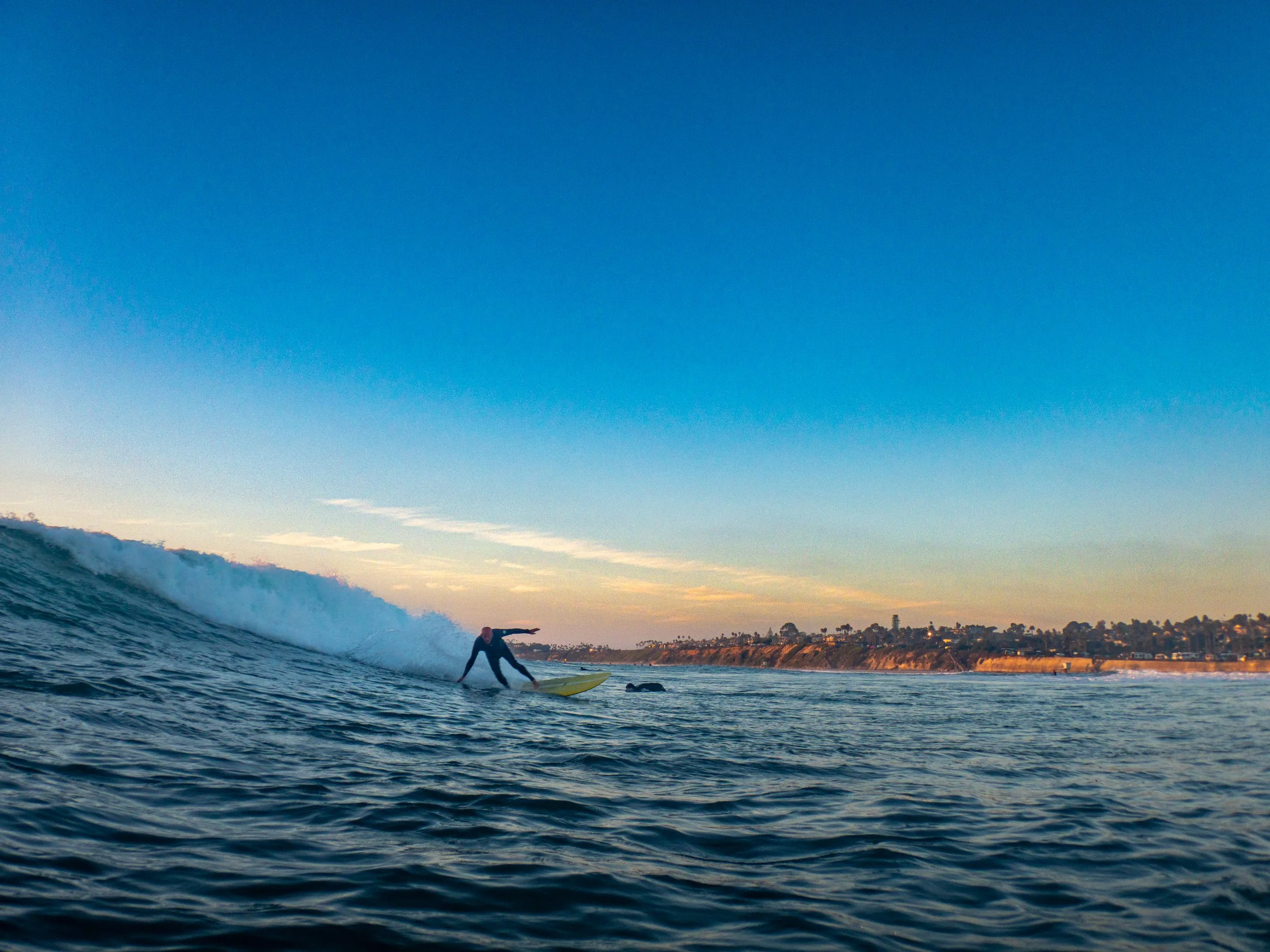 Surfing in the ocean at sunset with a wave and a sandy coastline in the background.