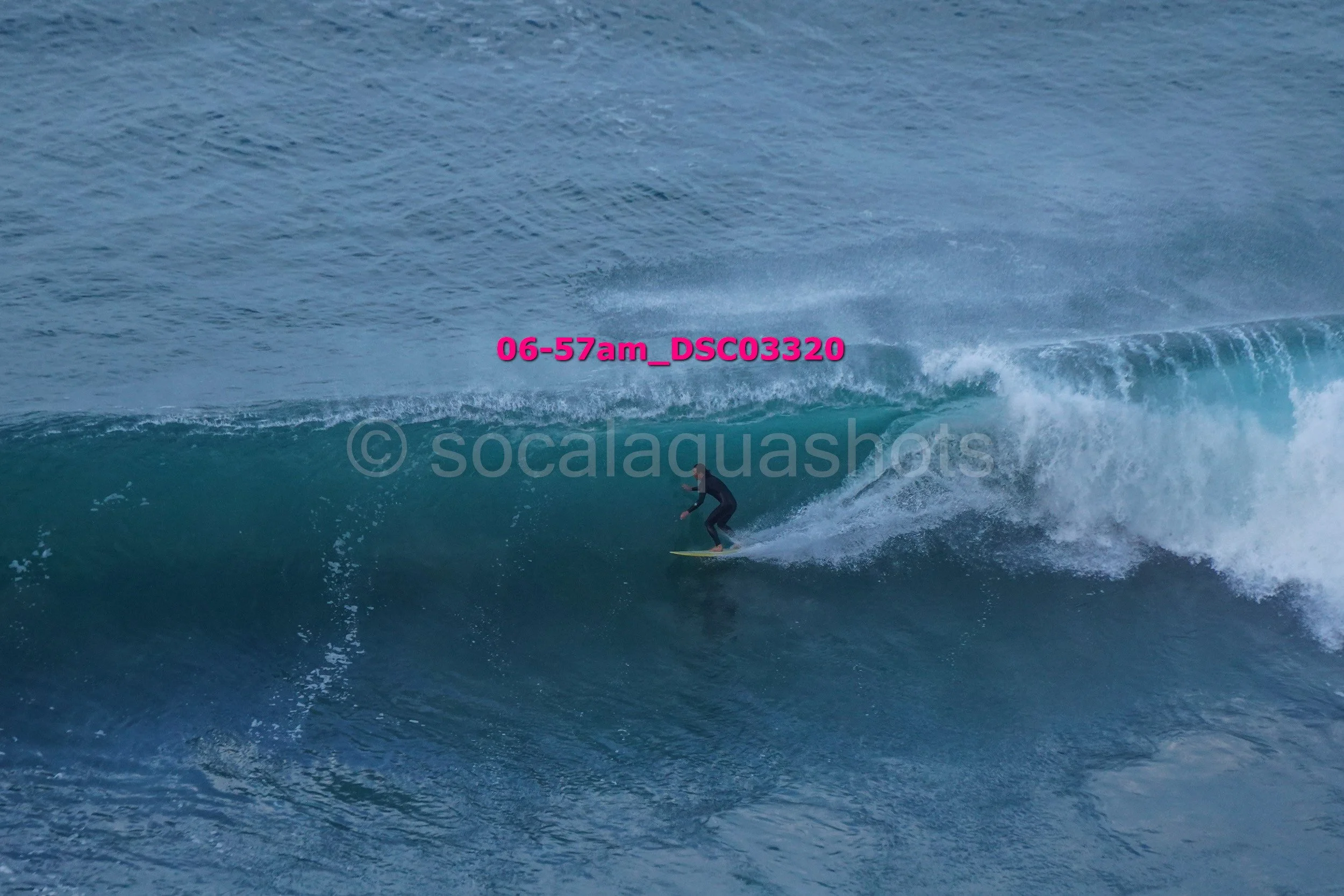 A person surfing on a wave in the ocean, wearing a black wetsuit and standing on a yellow surfboard.