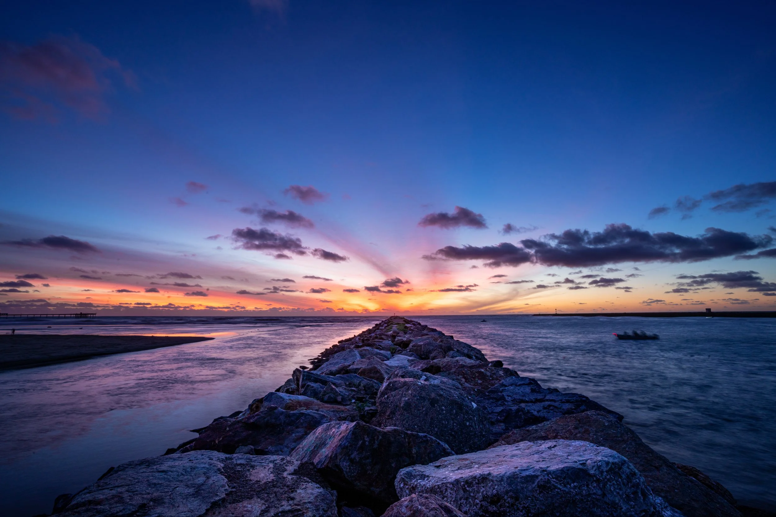 Sunset over the ocean with colorful sky, clouds, a rocky pier extending into the water, and a boat on the right side.