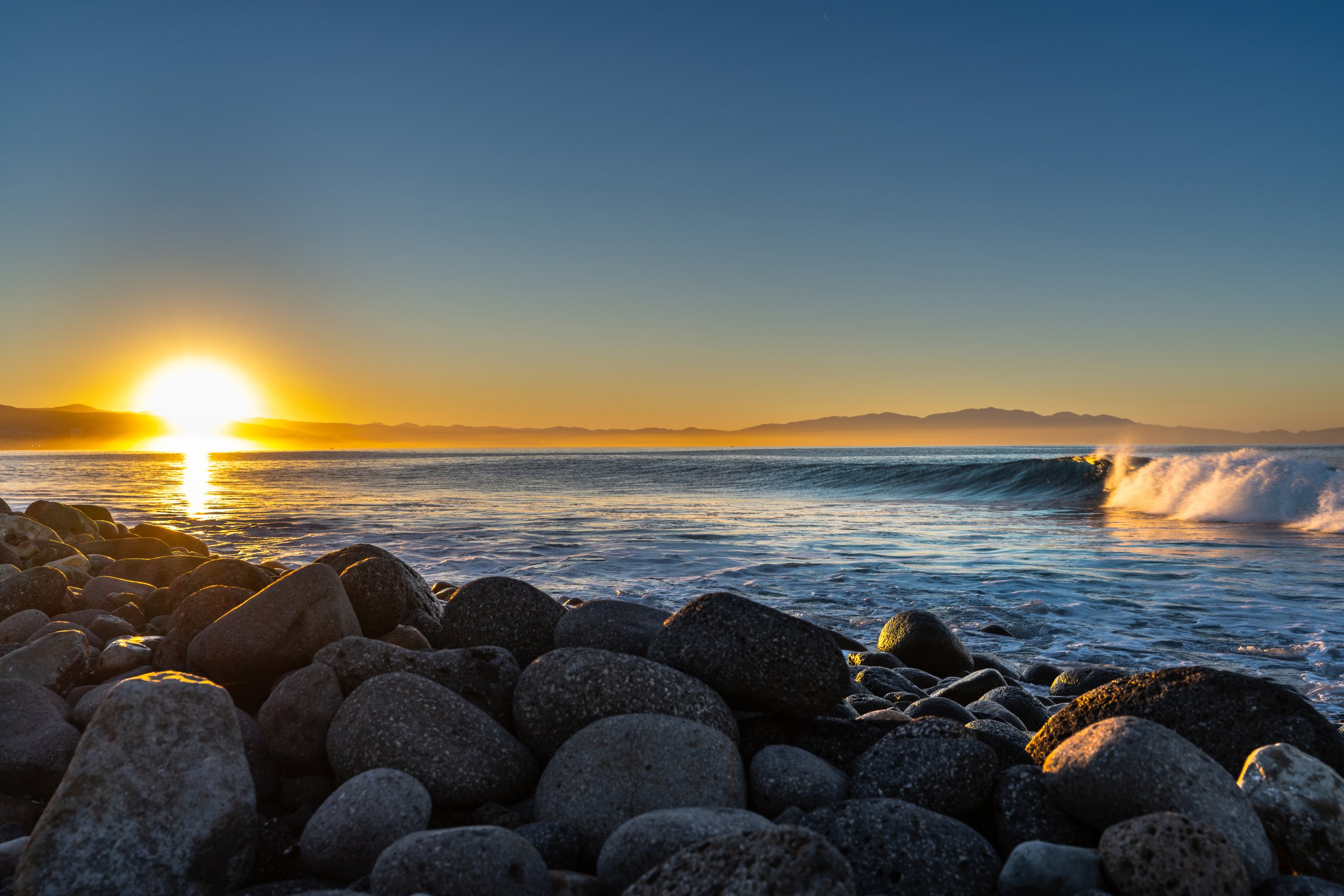 Sunset over the ocean with waves crashing on a rocky shoreline, mountains in the distance, and a clear sky.