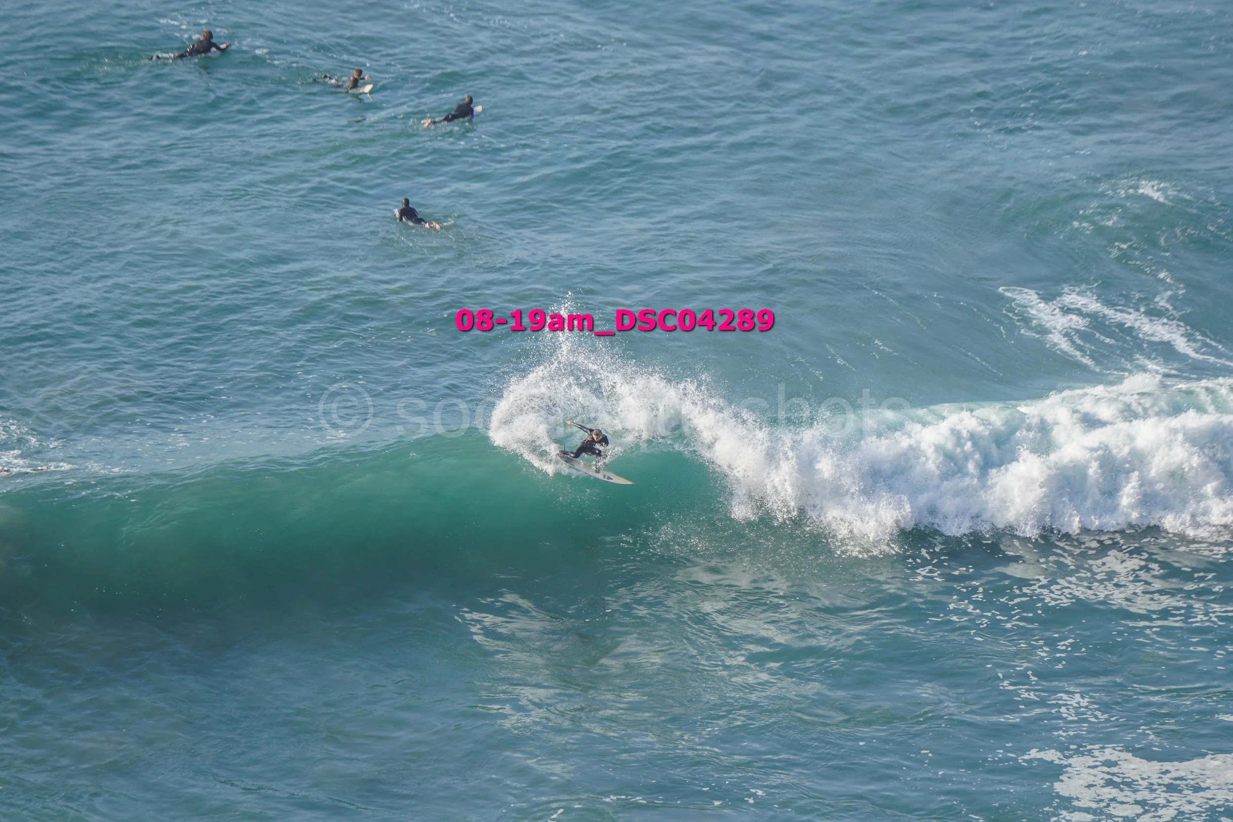 Surfer riding a wave in the ocean with several people swimming in the background.
