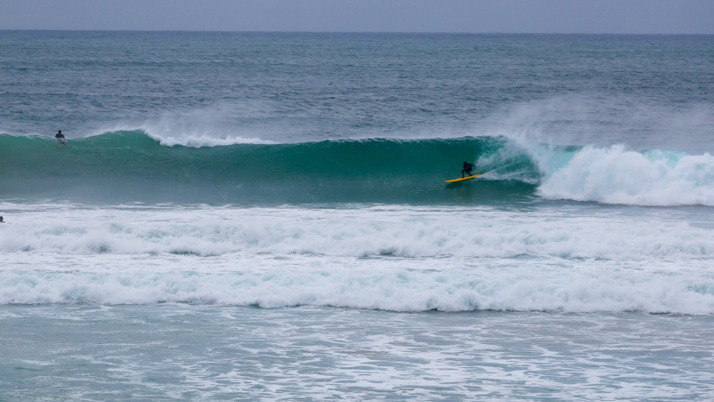 Surfer riding a wave on a yellow surfboard in the ocean, with another surfer in the background.