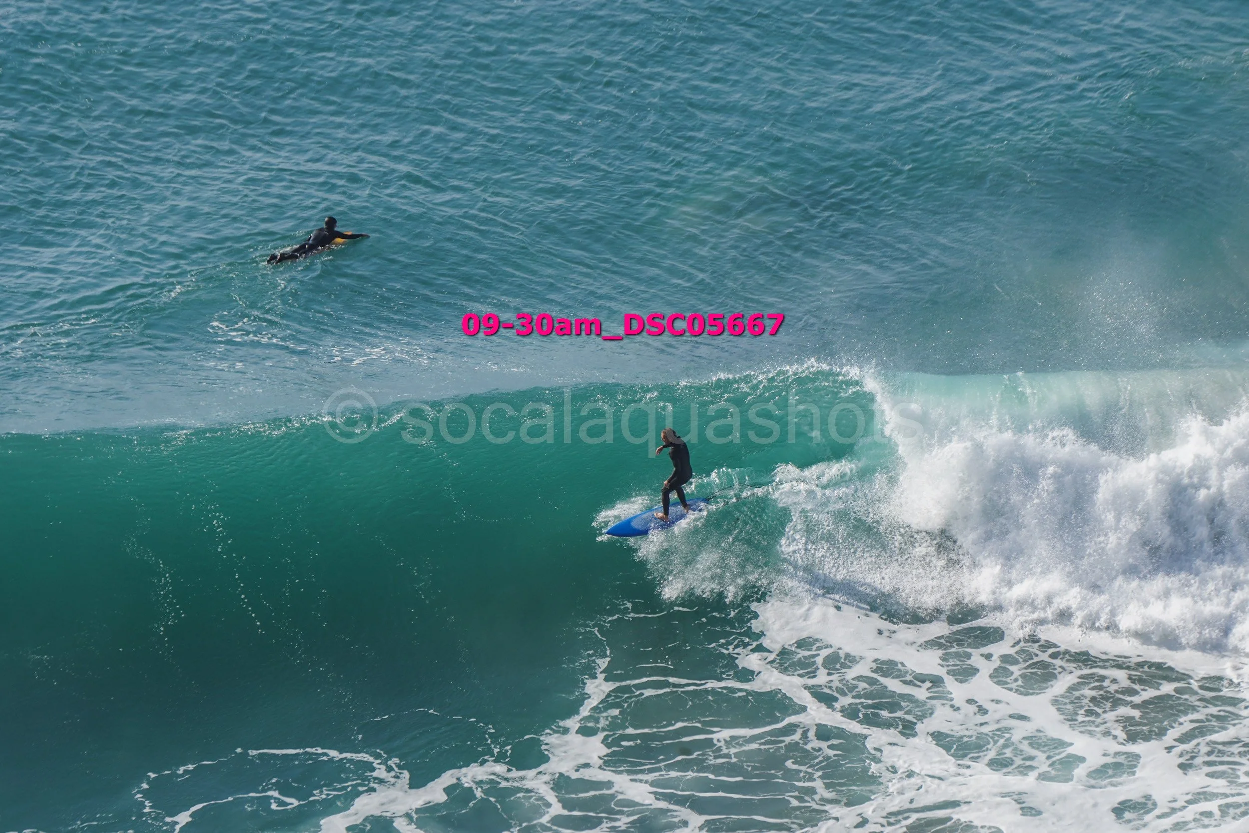 A surfer rides a large wave while another surfer paddles out in the ocean.