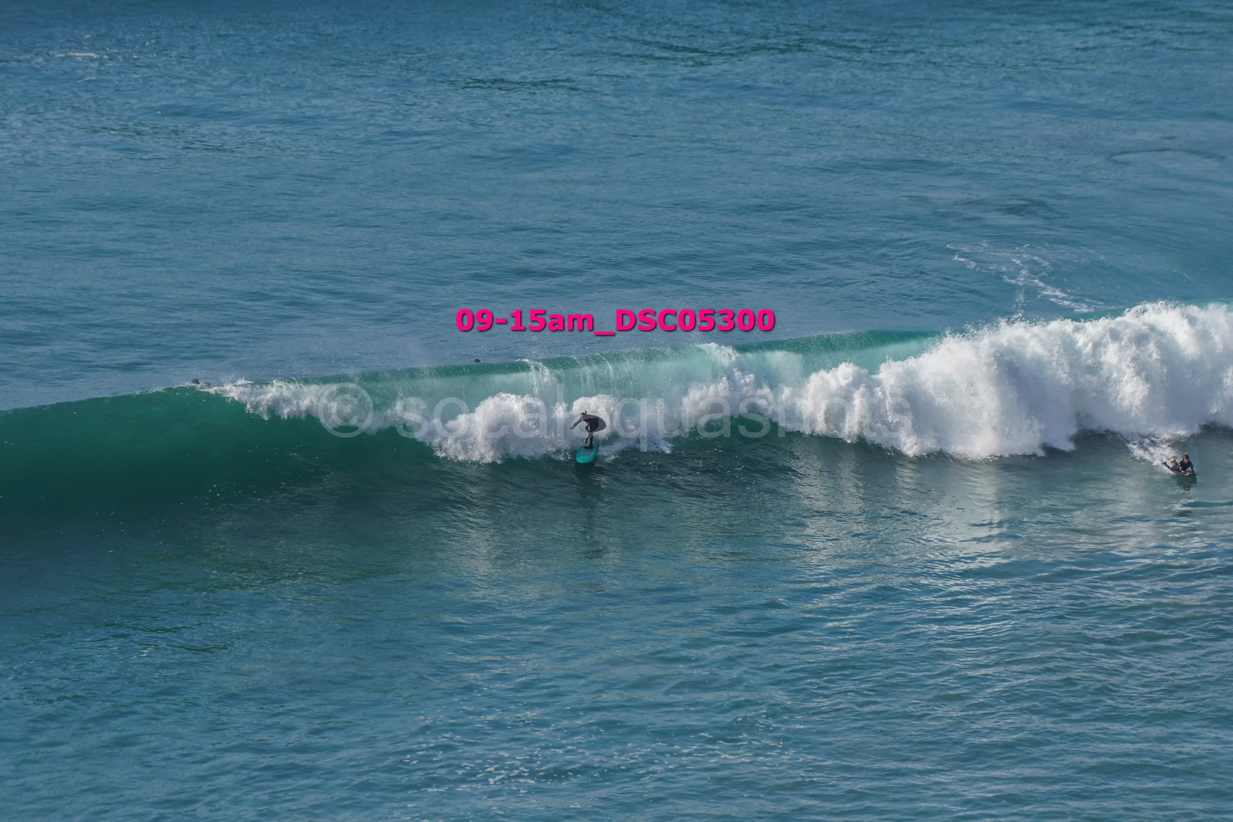 Surfer riding a wave in the ocean with another surfer on a surfboard nearby, blue water, no visible land.