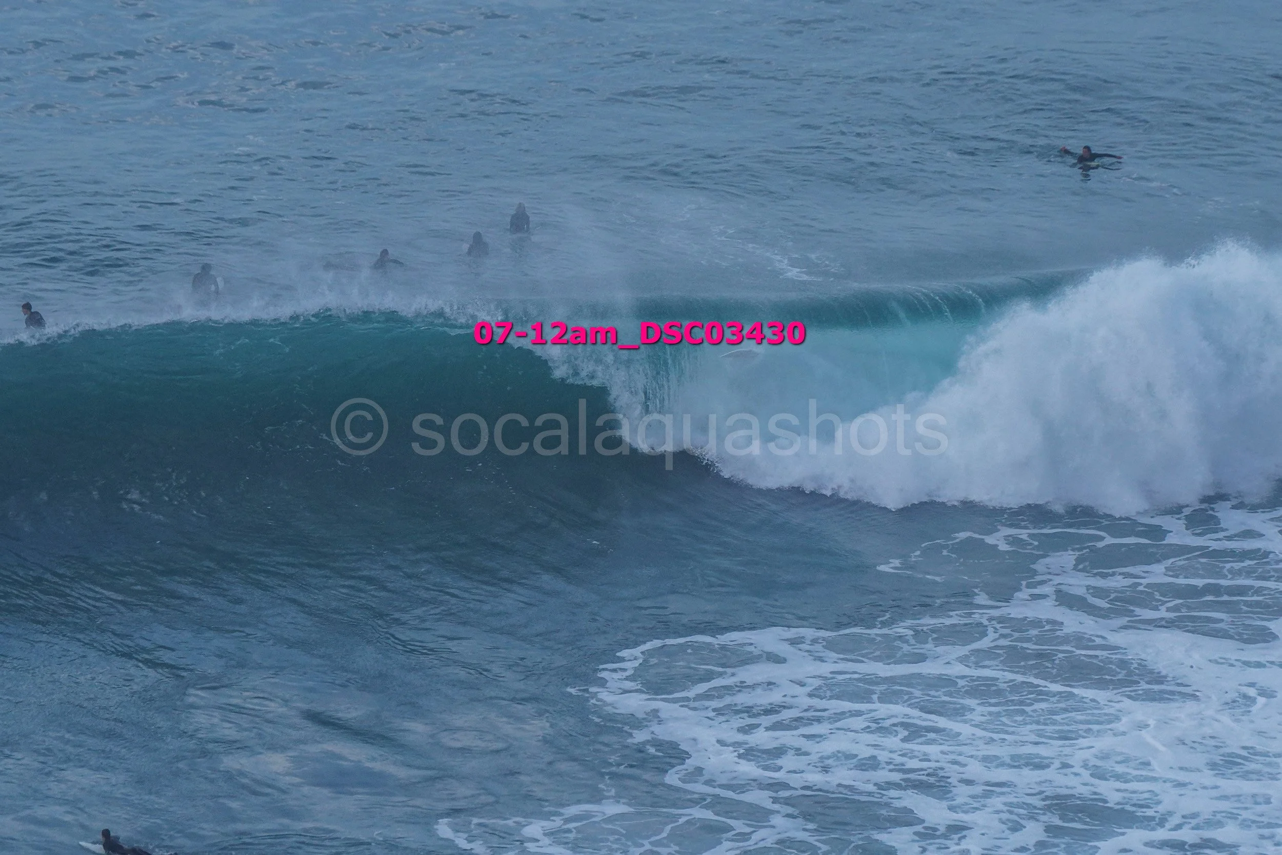 A group of surfers in the ocean, some riding a wave, others floating or waiting, with water surf and foam.