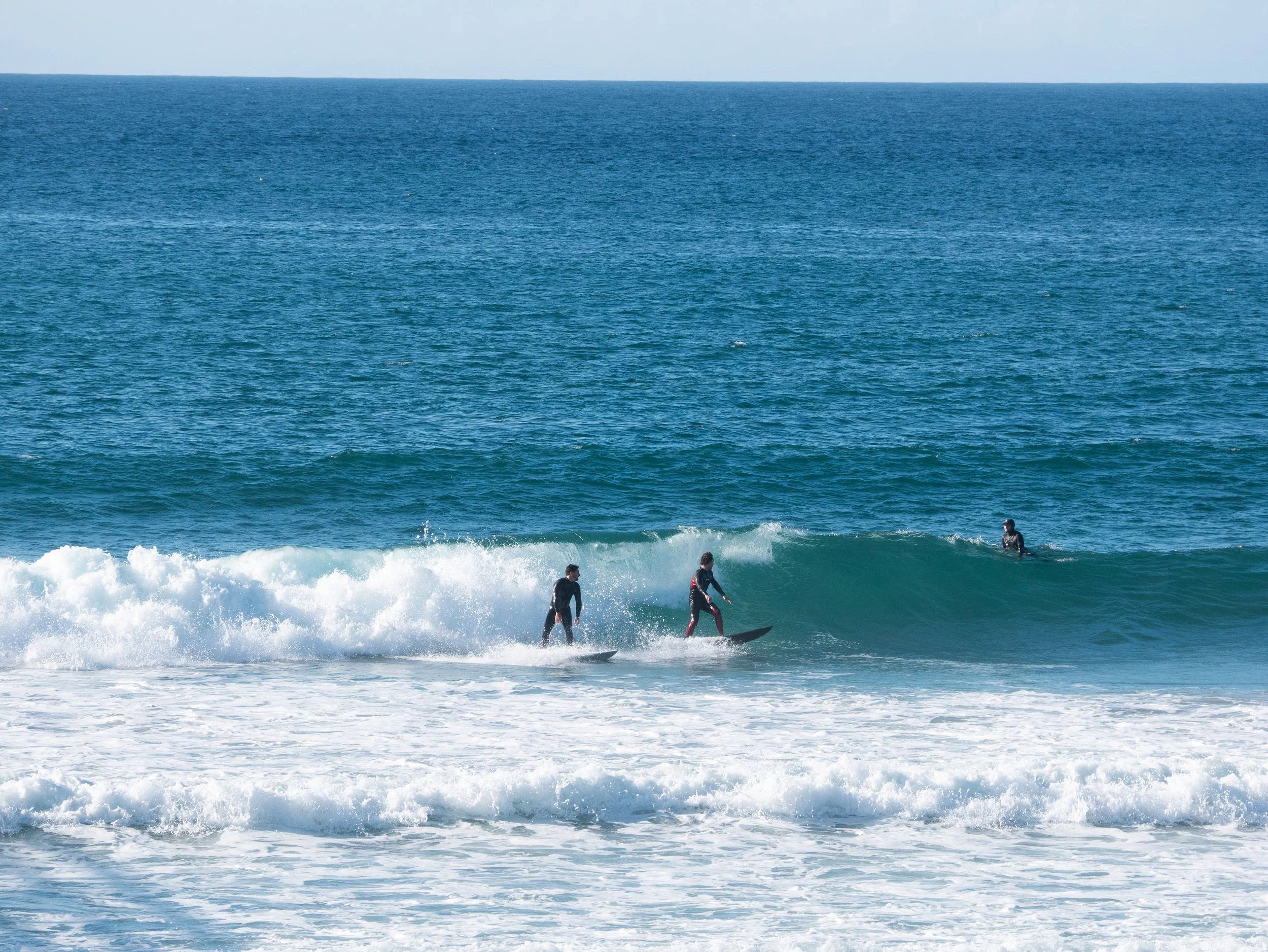 Three surfers riding waves at the beach with clear blue water.