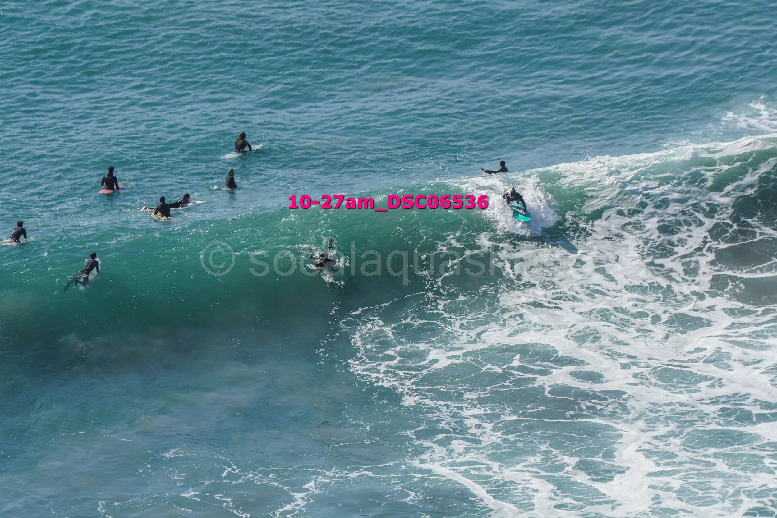 Surfers waiting in the ocean, some riding a wave, with clear blue water.