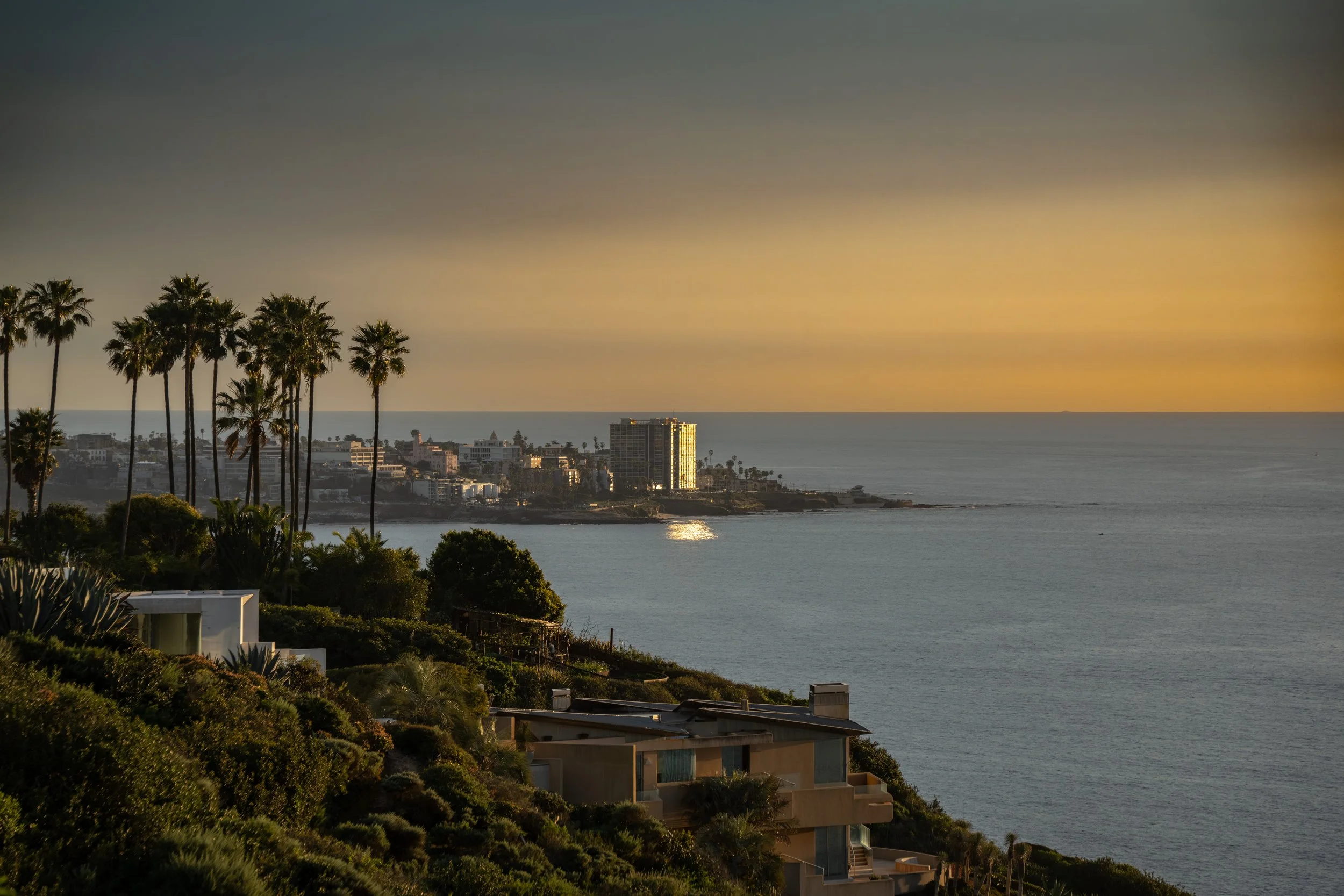 A coastal cityscape at sunset with palm trees, residential buildings in the foreground, and a large hotel or apartment building on a small peninsula by the ocean in the background.