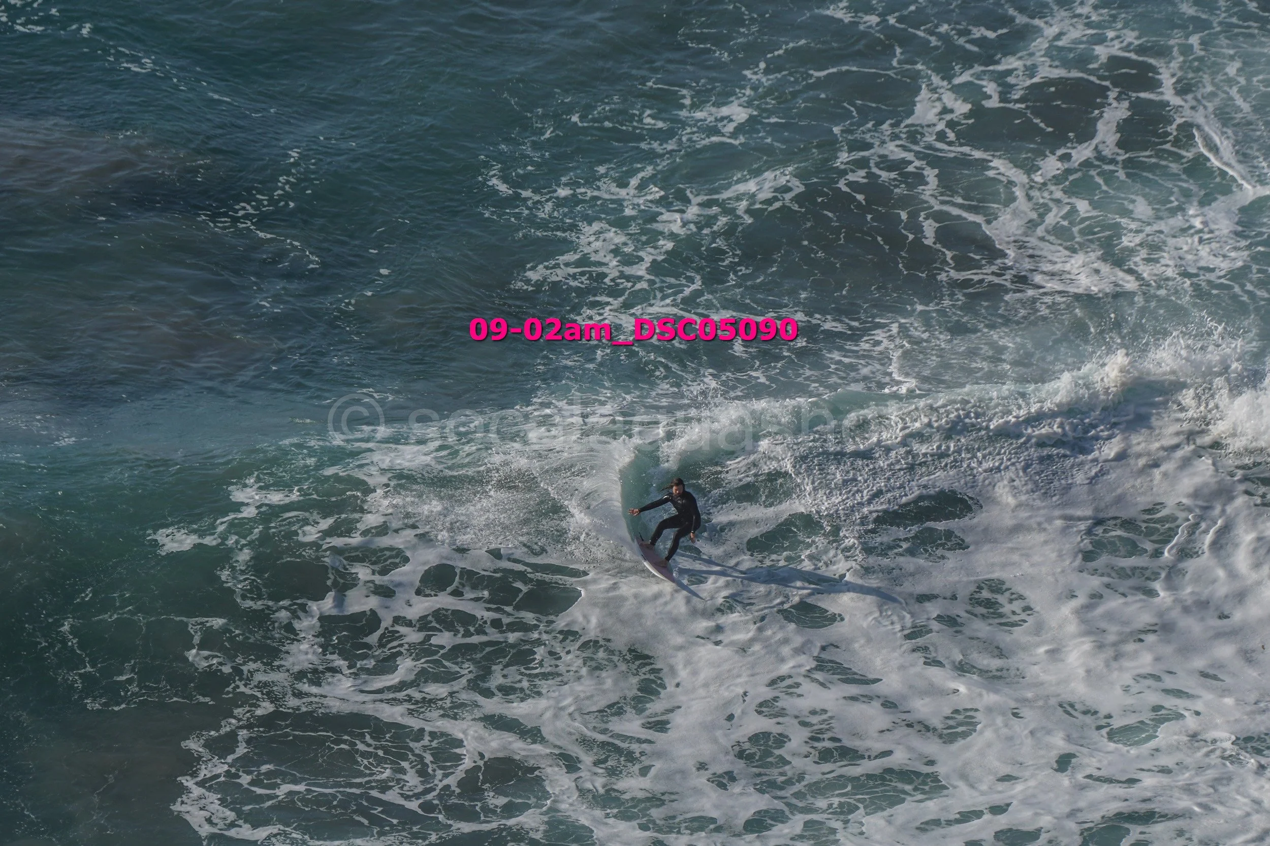 A person surfing on a wave in the ocean, wearing a black wetsuit, with the water creating white foam around them.
