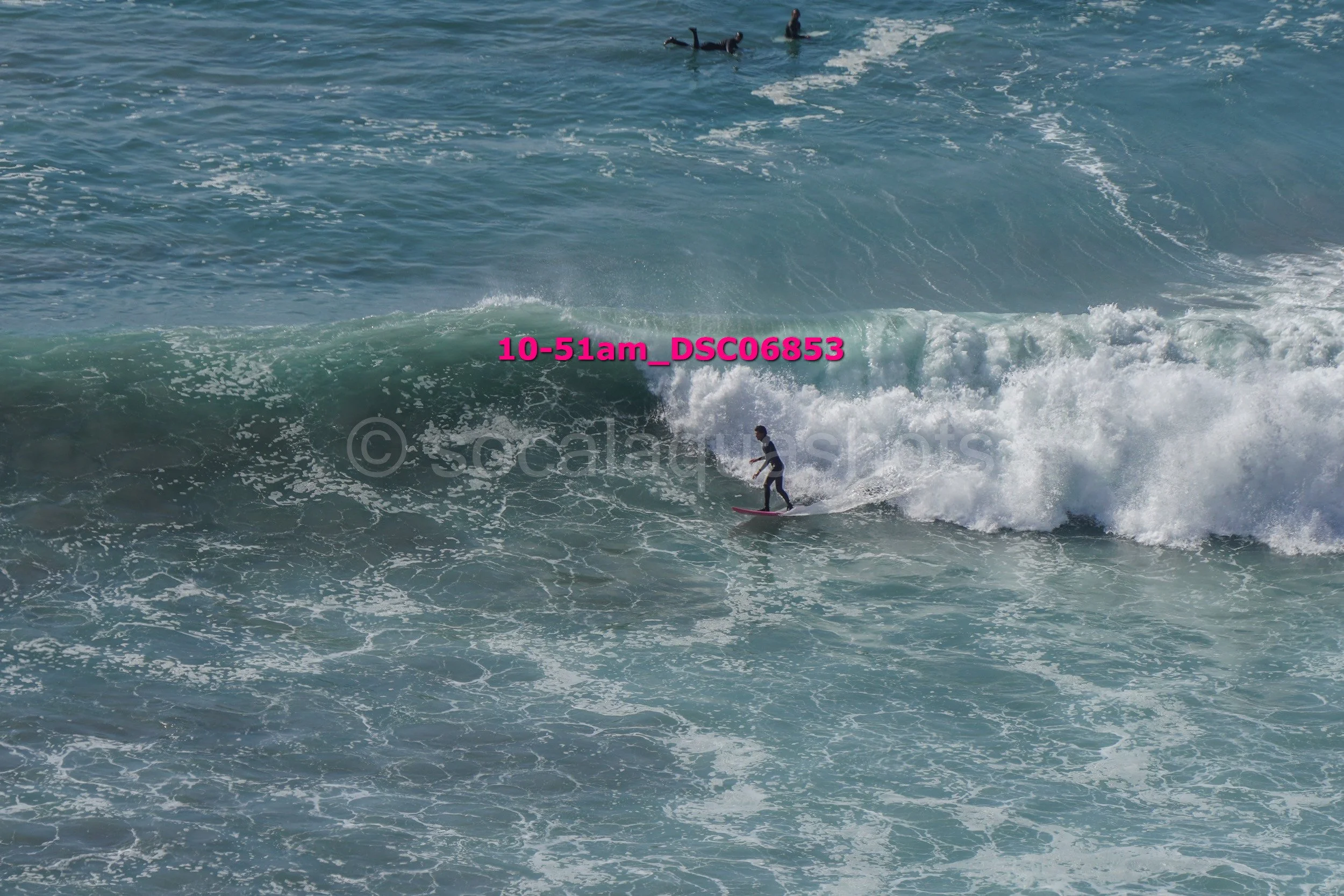 A person surfing on a small wave in the ocean, wearing a black wetsuit, with two other surfers in the distance on the water.