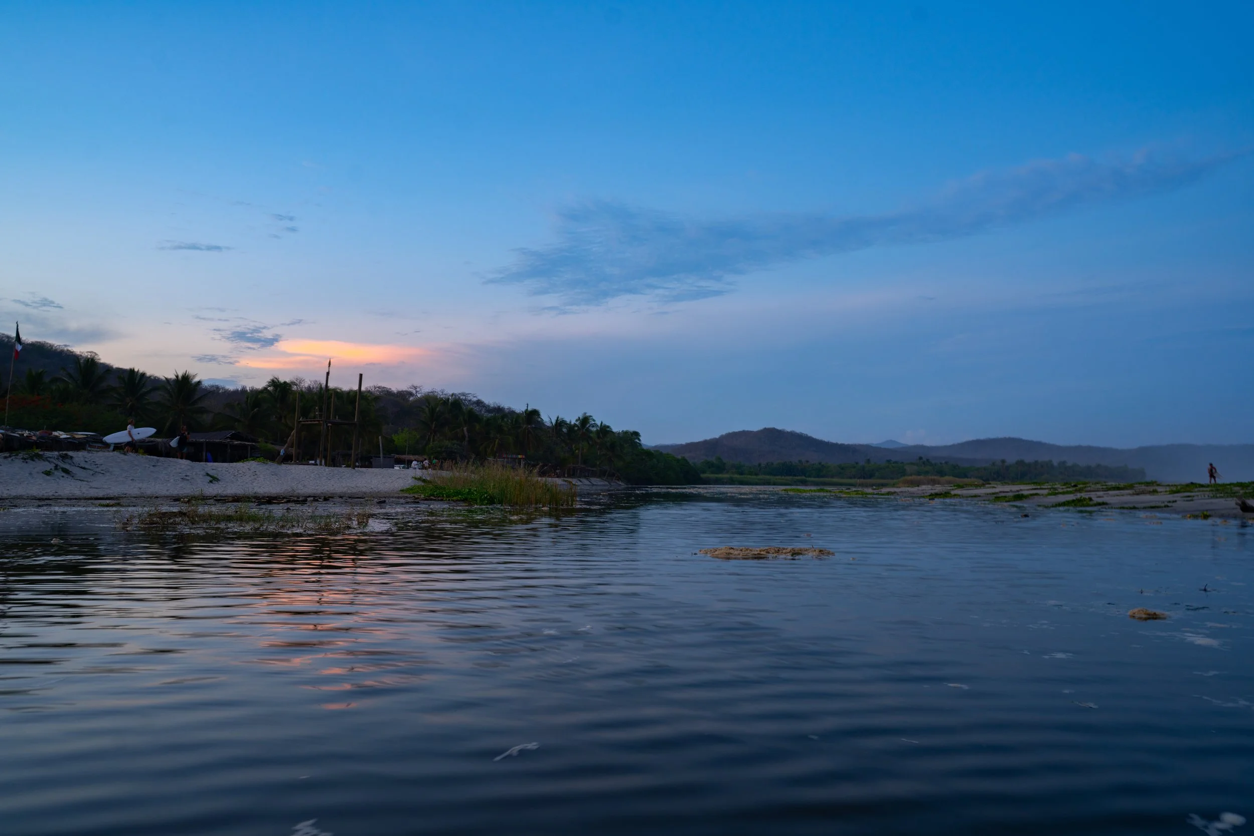 A peaceful river scene at dusk with calm water, a distant shoreline with trees, palm trees, and a small structure, under a sky with blue and orange hues.
