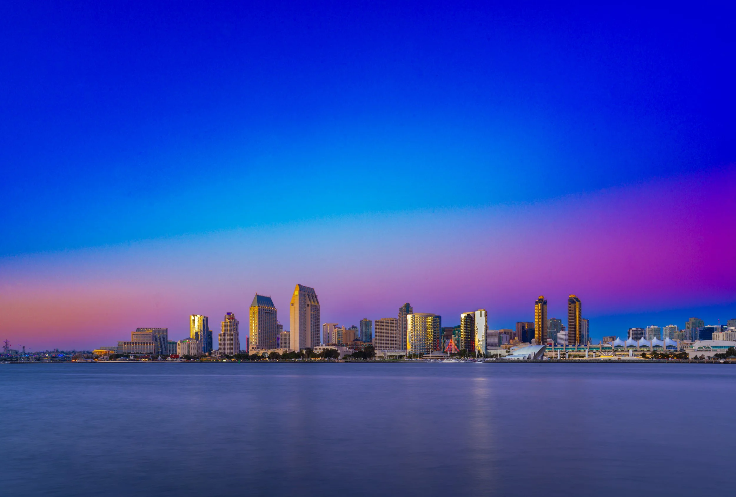 Night view of a city skyline reflecting on the water, with a colorful sky transitioning from pink to purple to blue.