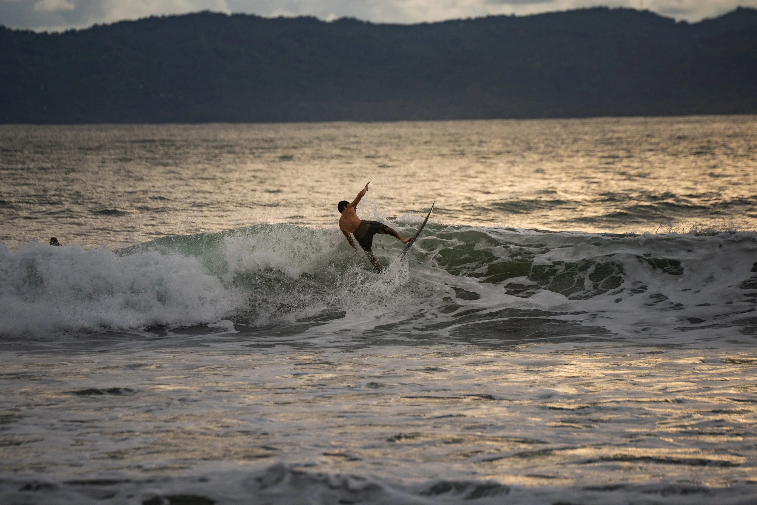 A person surfing on a wave in the ocean at sunset, with mountains in the background.