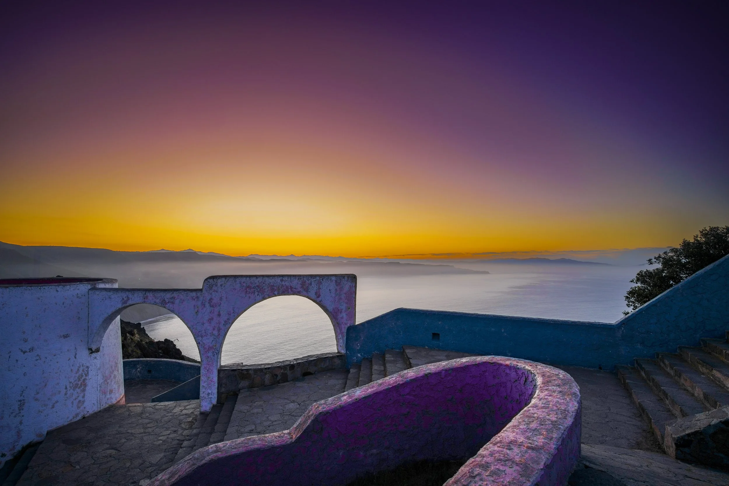 Colorful sunset over the ocean, with a white and blue architectural structure in the foreground, including staircases and archways.