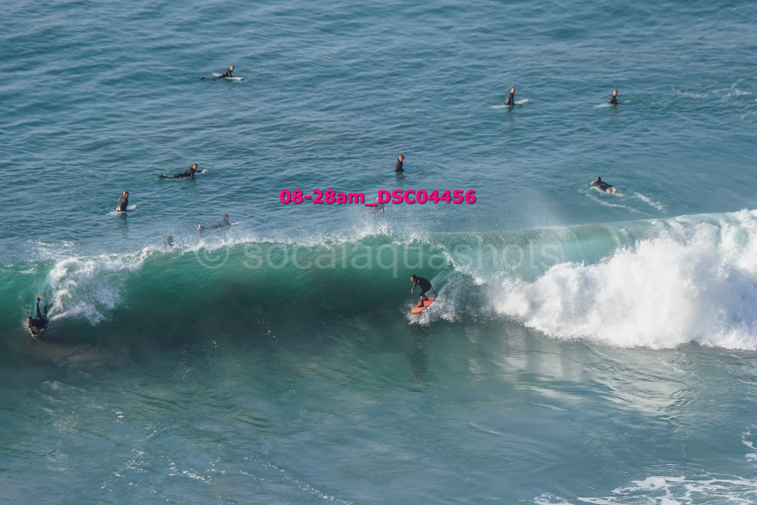 Surfer riding a wave while multiple surfers in wetsuits wait in the water nearby.