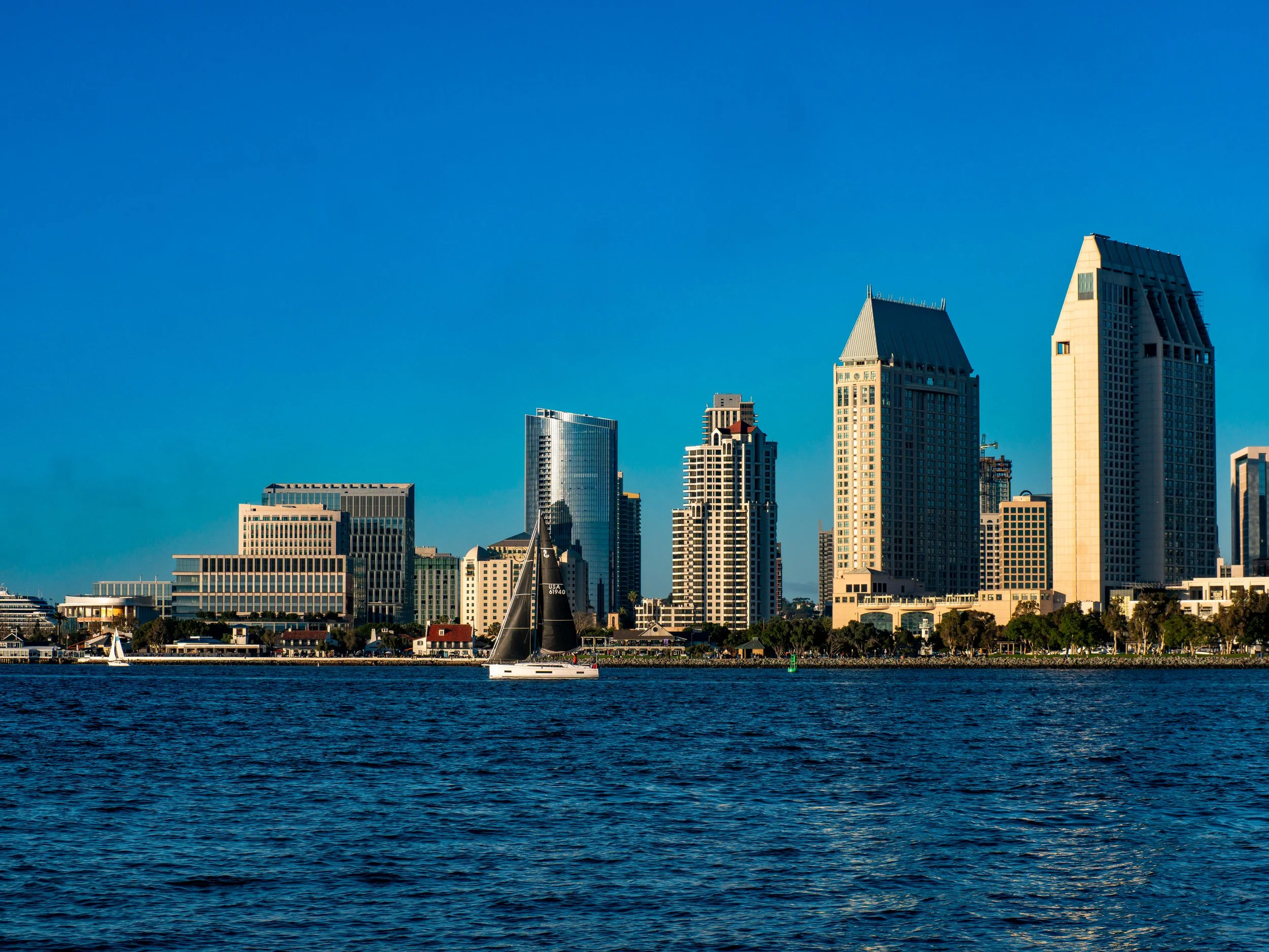 A city skyline with tall modern buildings along the waterfront, with a sailboat on the water in the foreground and a clear blue sky overhead.