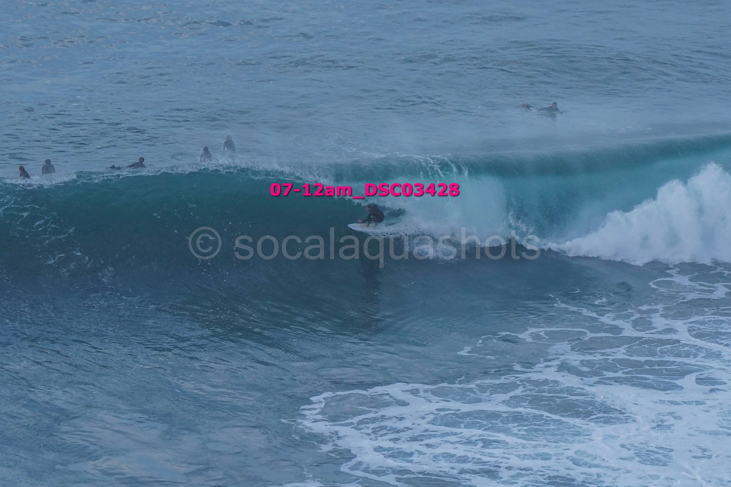 A person surfing on a large wave with several people in the water in the background.