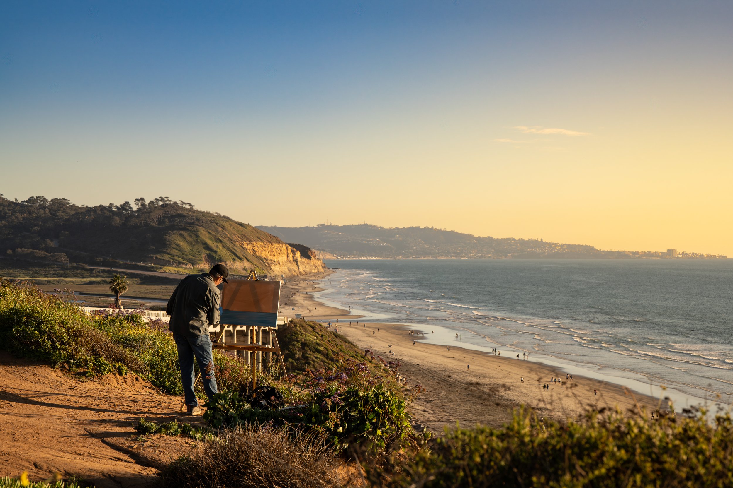 An artist painting on a canvas outdoors at a coastal viewpoint during sunset, overlooking a beach with ocean waves and cliffs in the background.