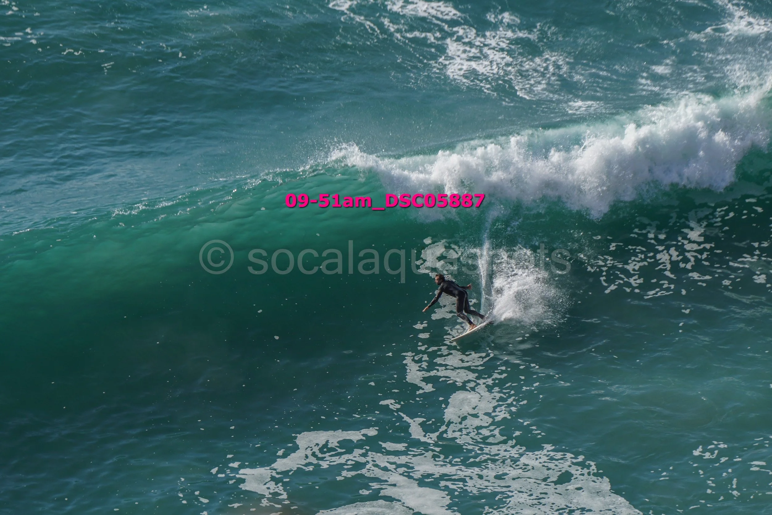 A surfer riding a large ocean wave with white foam at the crest, wearing a black wetsuit.