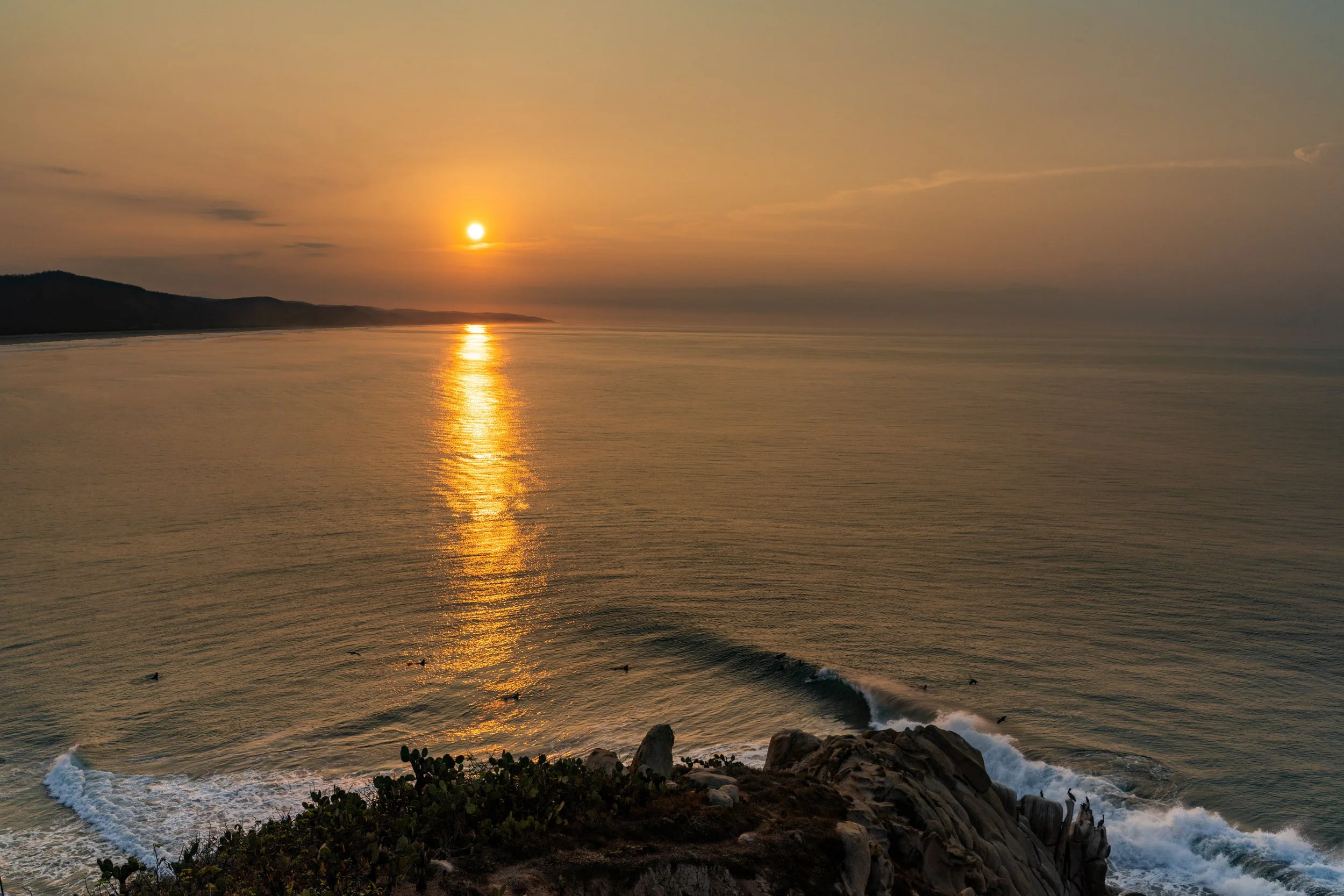 Sunset over the ocean with orange and yellow sky, reflective water, rocky coastline, and small waves crashing against rocks.
