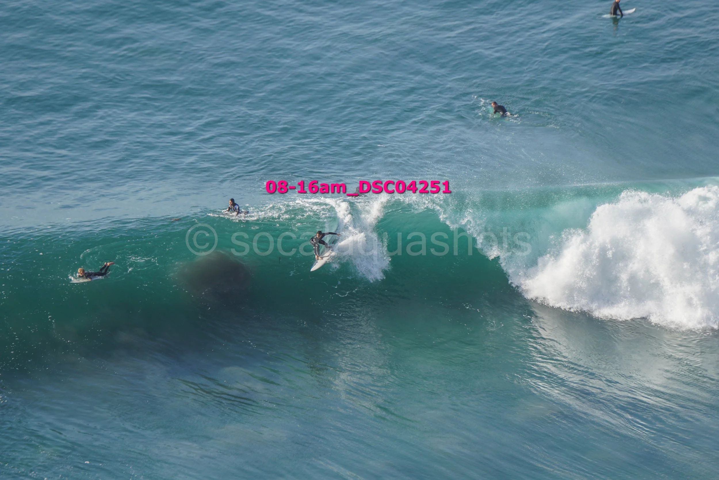 Surfers riding and waiting for waves in the ocean, with one surfer actively riding a wave and others floating or paddling nearby.