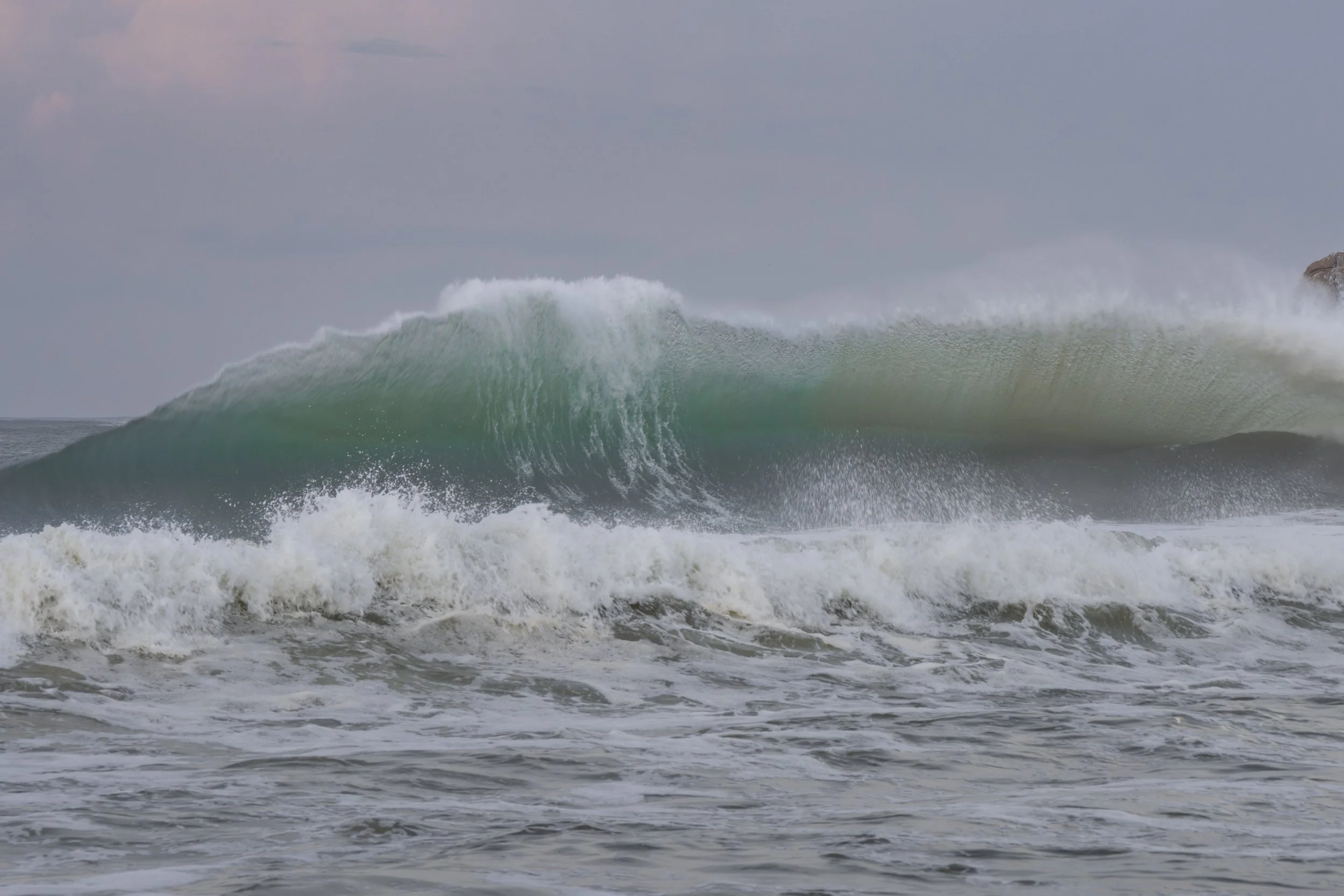 Ocean wave crashing, with a large wave cresting over and spray in the air during cloudy weather.