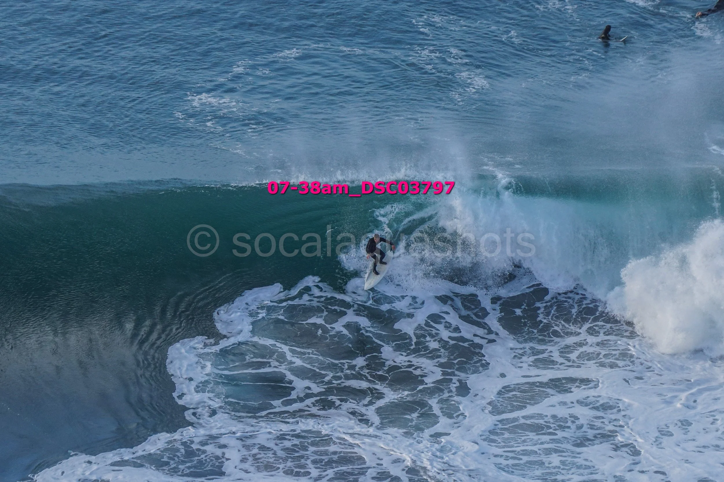 A person surfing on a large ocean wave with several other surfers visible in the water in the background.
