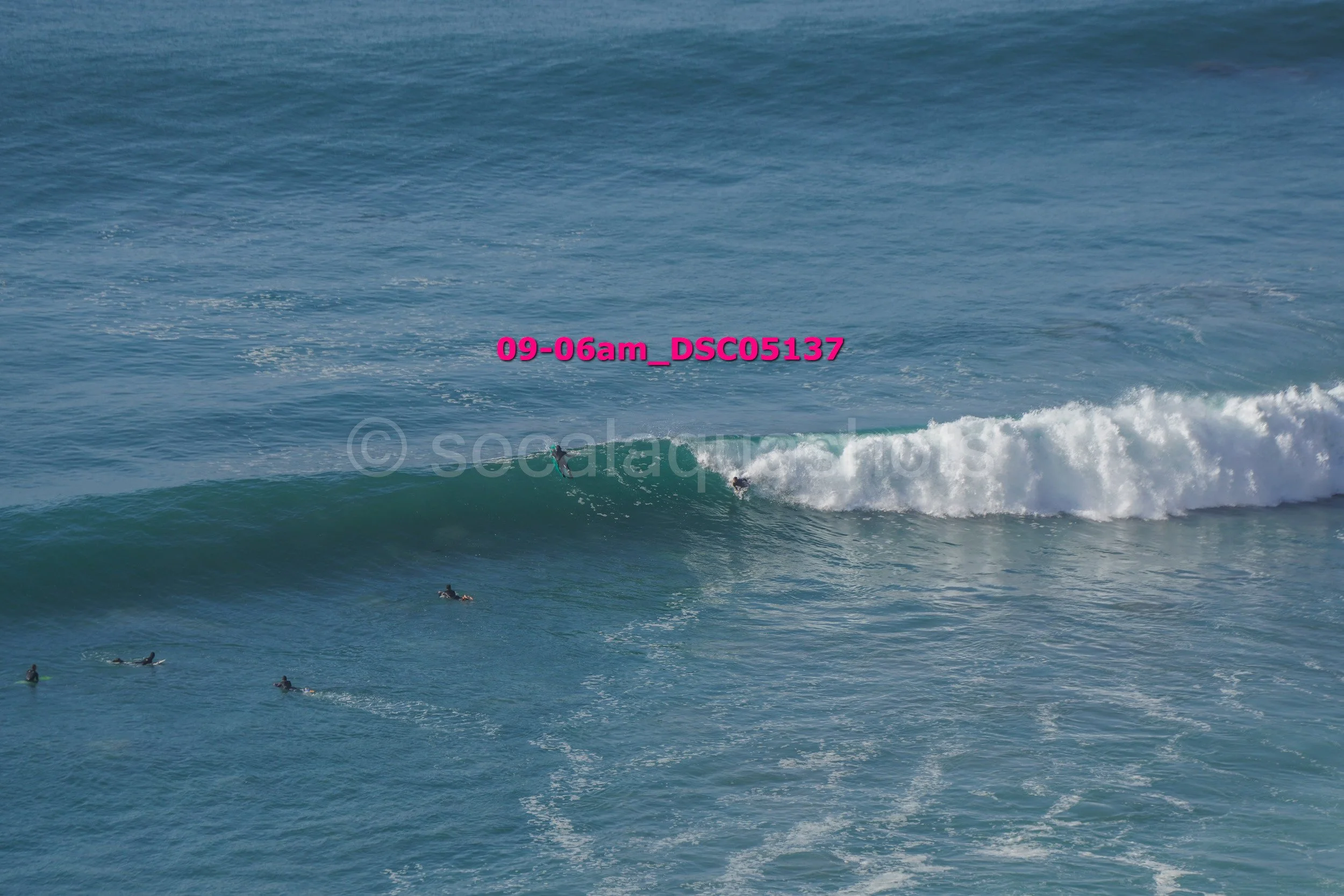 Surfers in the ocean, some riding a wave while others wait in the water, with blue water and white surf.