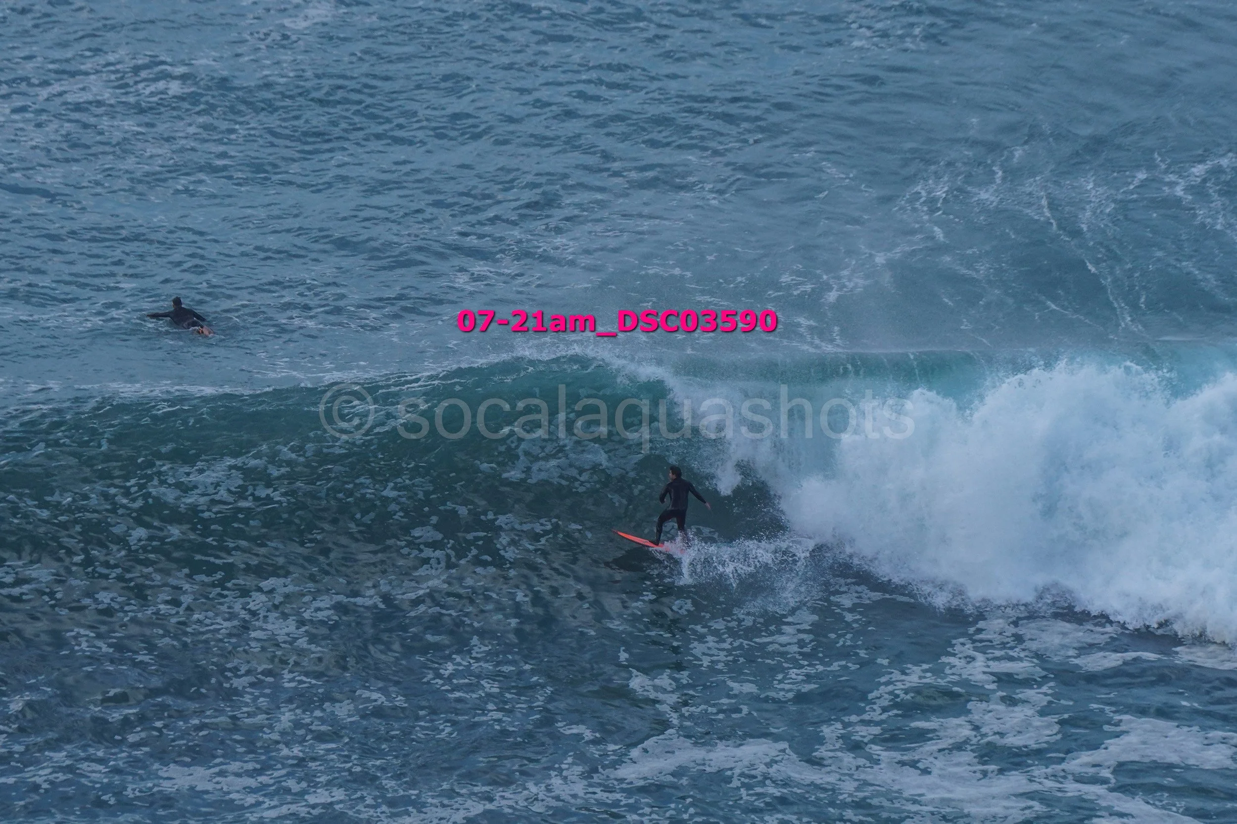 Two surfers riding a wave in the ocean, one standing on a surfboard and the other swimming nearby.