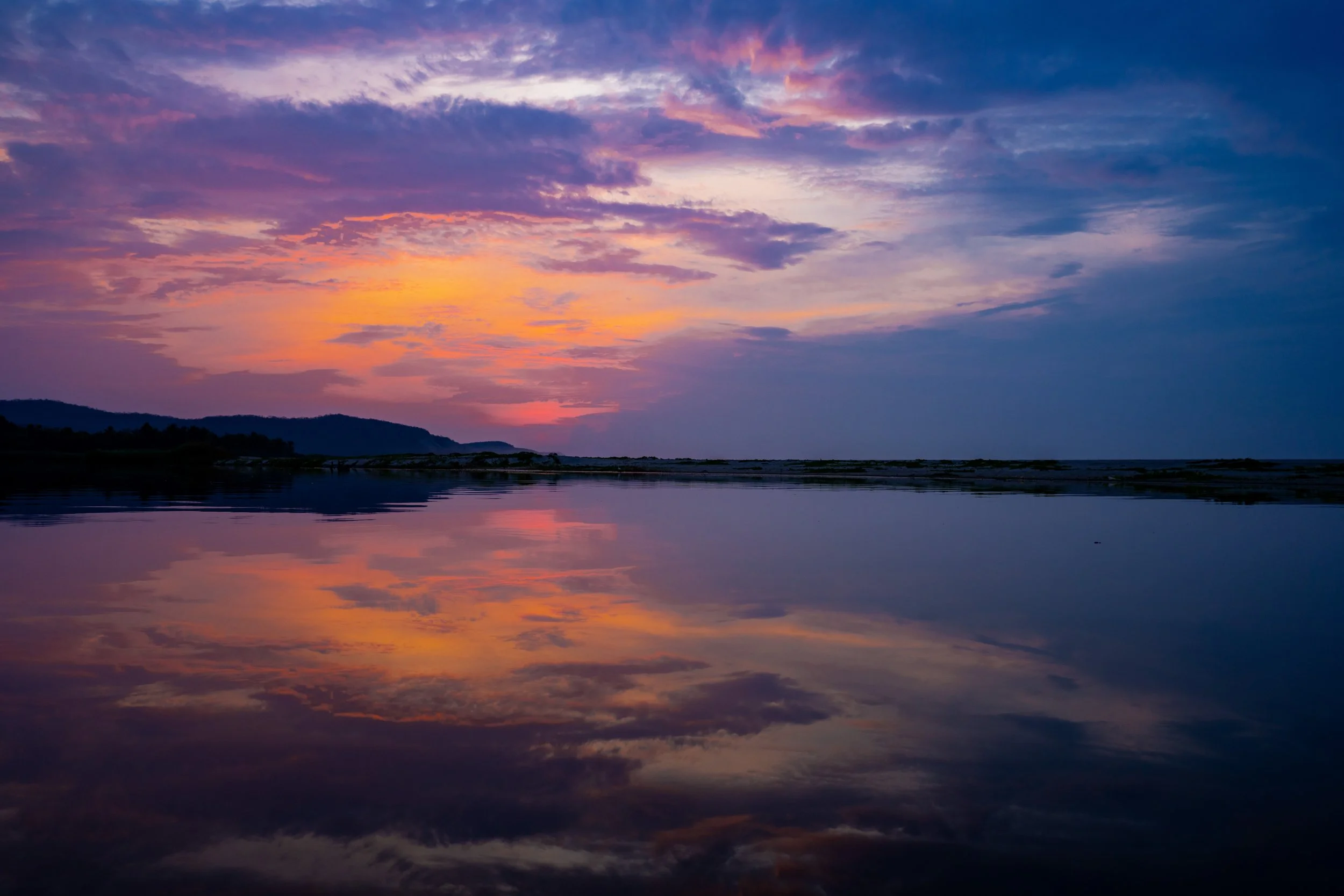 A colorful sunset over a calm body of water, with vibrant pink, purple, orange, and blue clouds reflected on the surface.