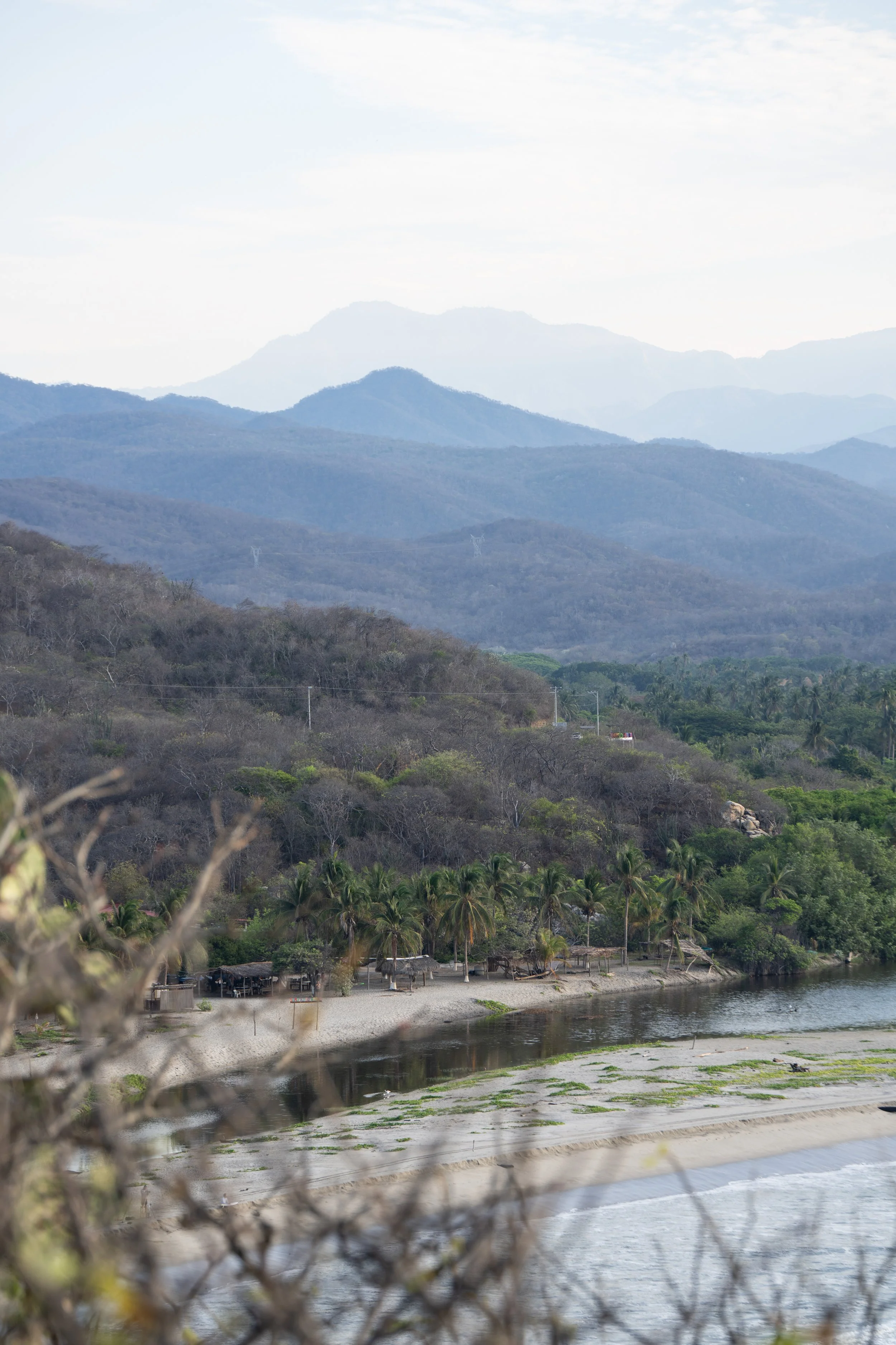 A scenic landscape with distant mountains, a river, and trees along the riverbank, with a small beach area and huts at the water's edge.