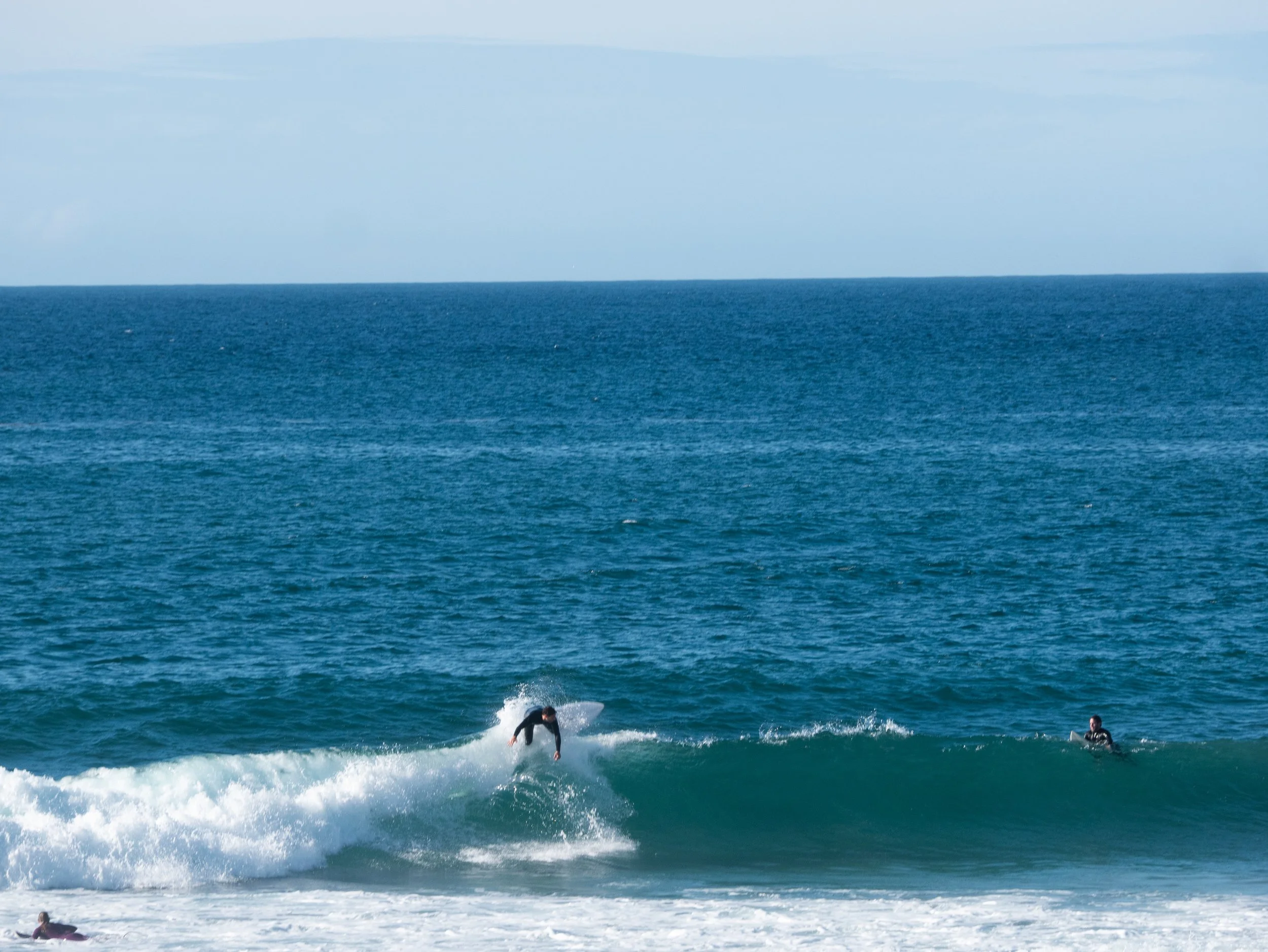 A person surfing on a small wave in the ocean, with a person in the water nearby, under a partly cloudy sky.