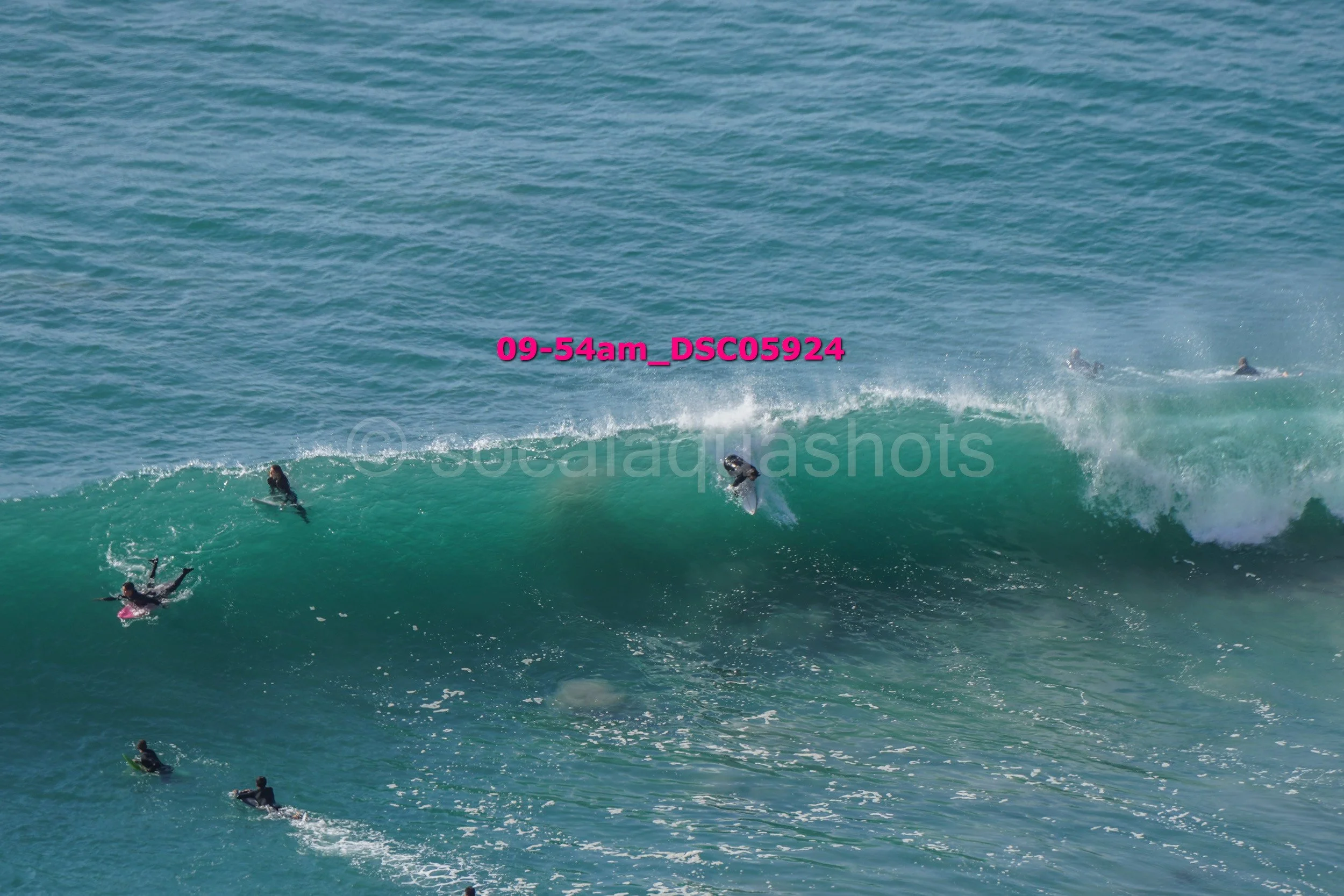 Several surfers riding and floating in the ocean waves, with a large wave breaking and surf spray visible.