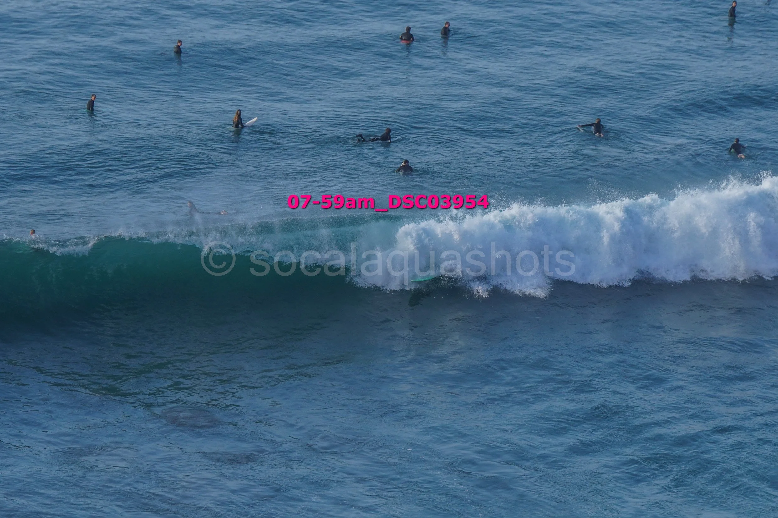 Surfers in the ocean, some sitting on surfboards and waiting, others riding a wave, with blue water and white foam.