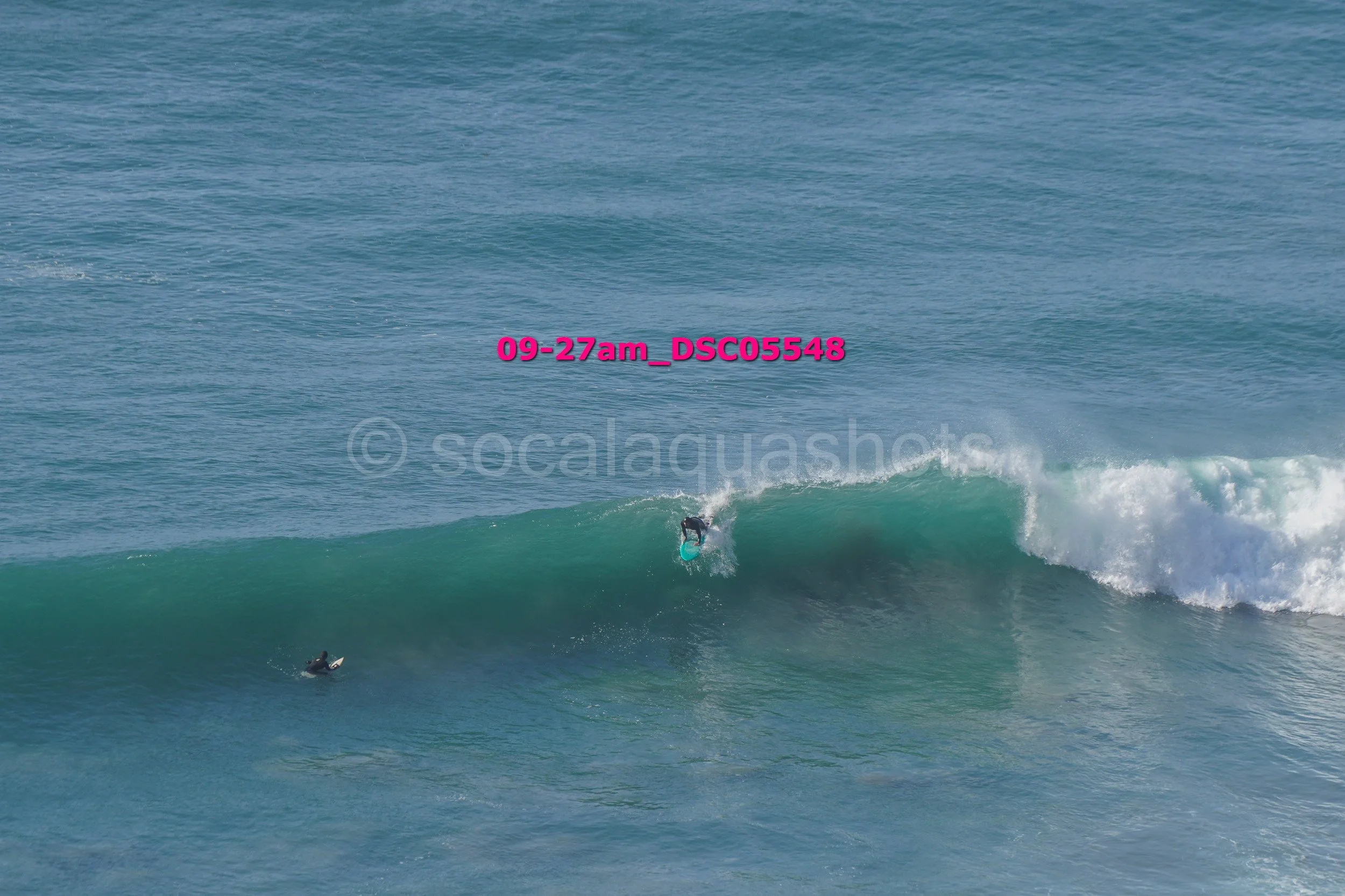 A person surfing a large wave in the ocean, with another surfer in the water watching.