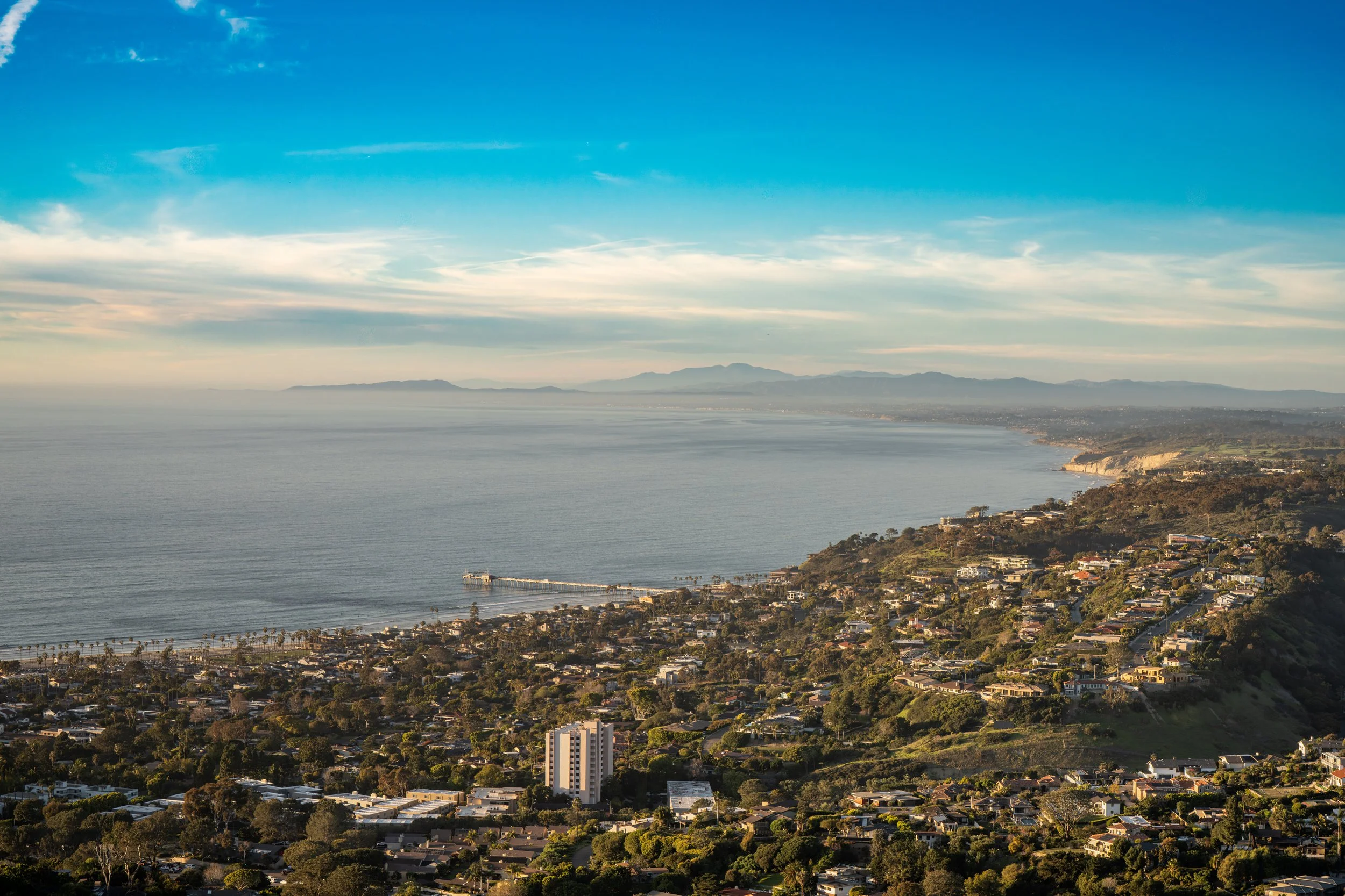 A coastal cityscape with residential houses on hills, a beach with a pier extending into calm ocean waters, and mountains in the distance under a partly cloudy sky.