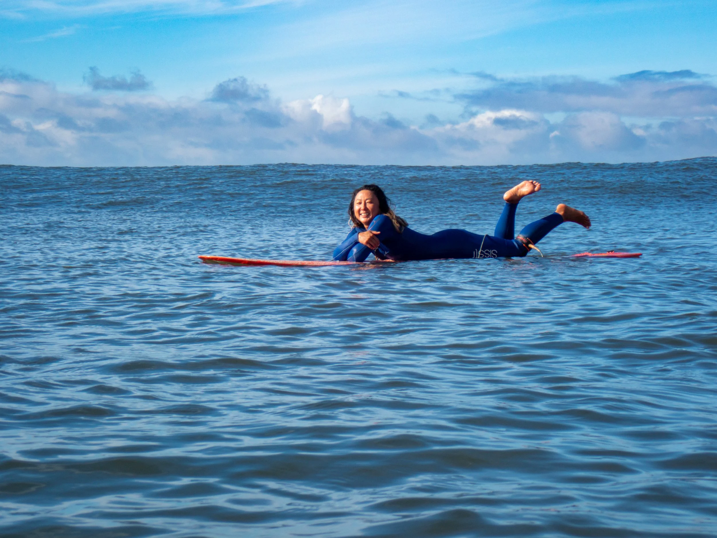 Woman in a wetsuit lying on a surfboard in the ocean, smiling, with a partly cloudy sky in the background.